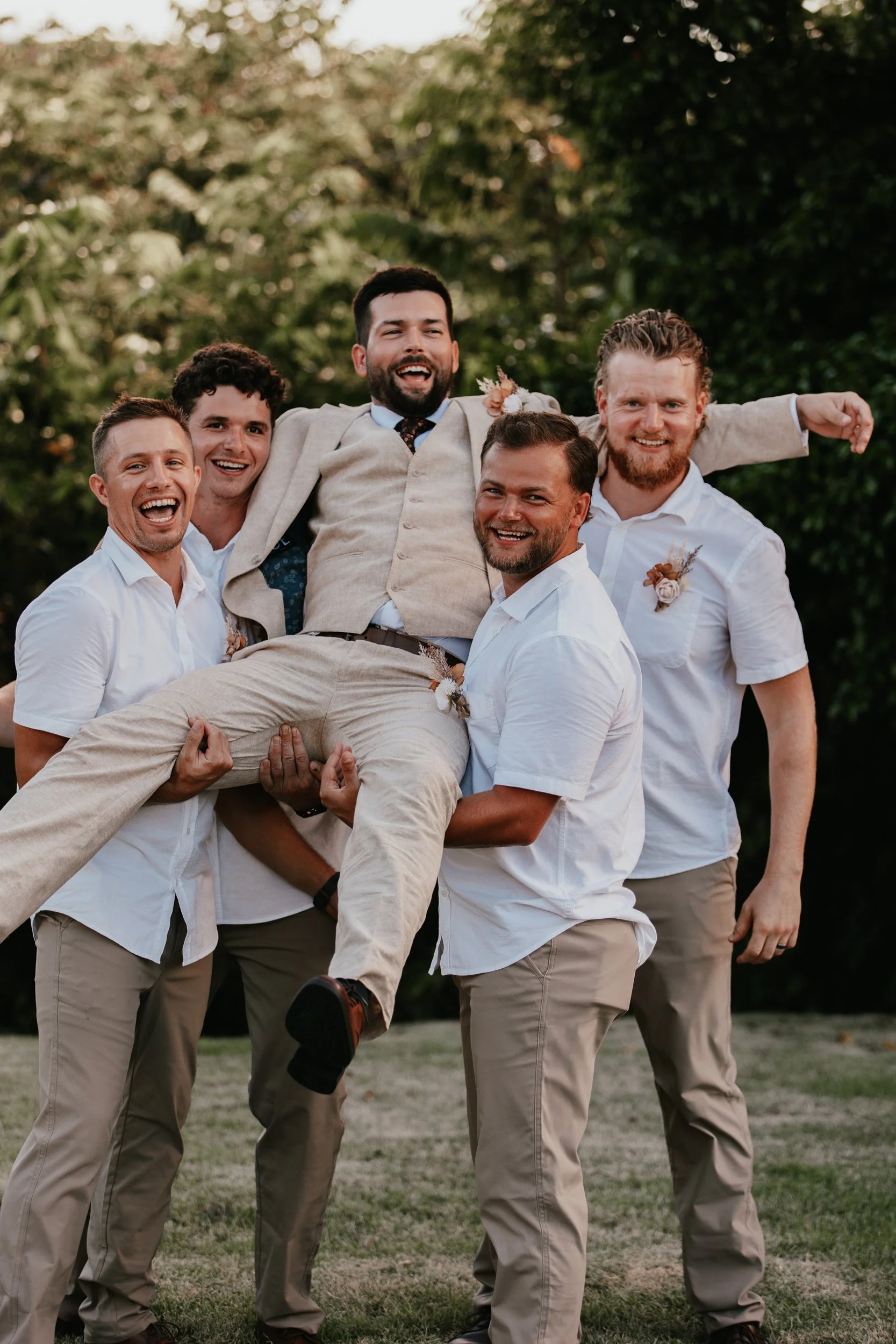 Group of men lifting a groom in a tan suit at an outdoor wedding, smiling and celebrating.