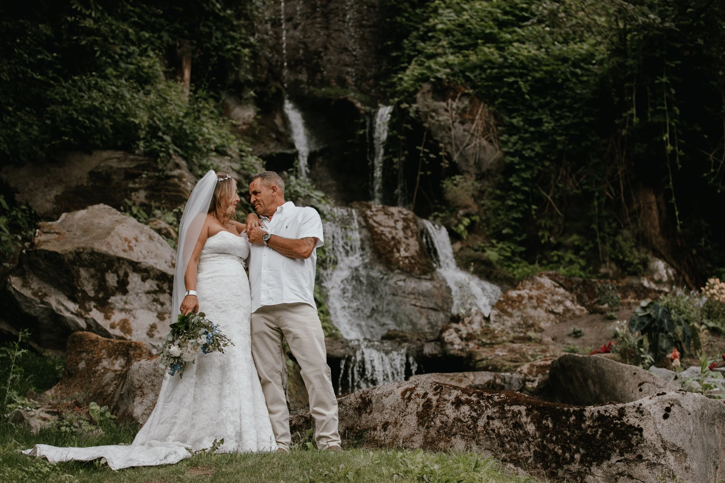 A bride in a white wedding gown and a groom in a white shirt and beige pants standing close together outdoors near a waterfall surrounded by rocks and lush greenery.
