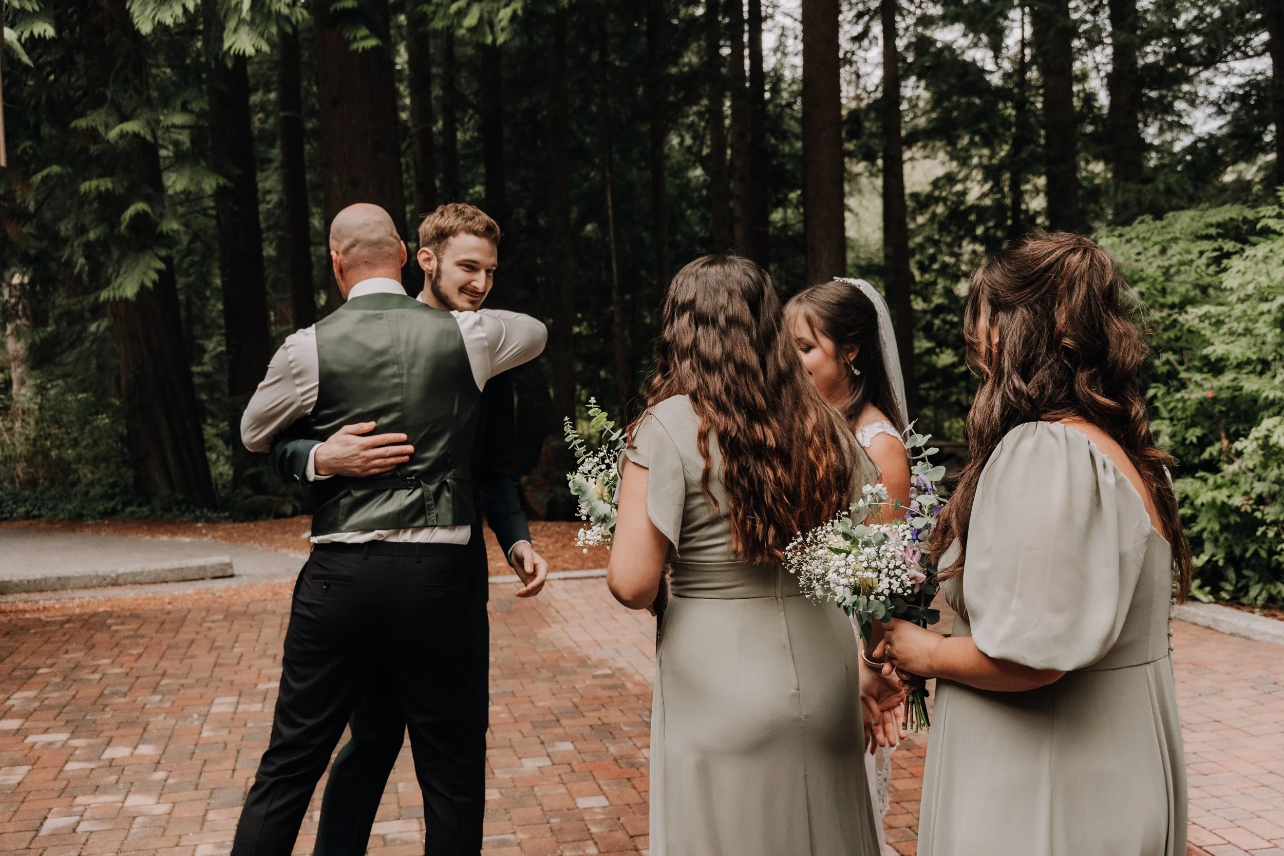 People celebrating a wedding outdoors, with two men hugging and two women holding bouquets of flowers, in a wooded setting.