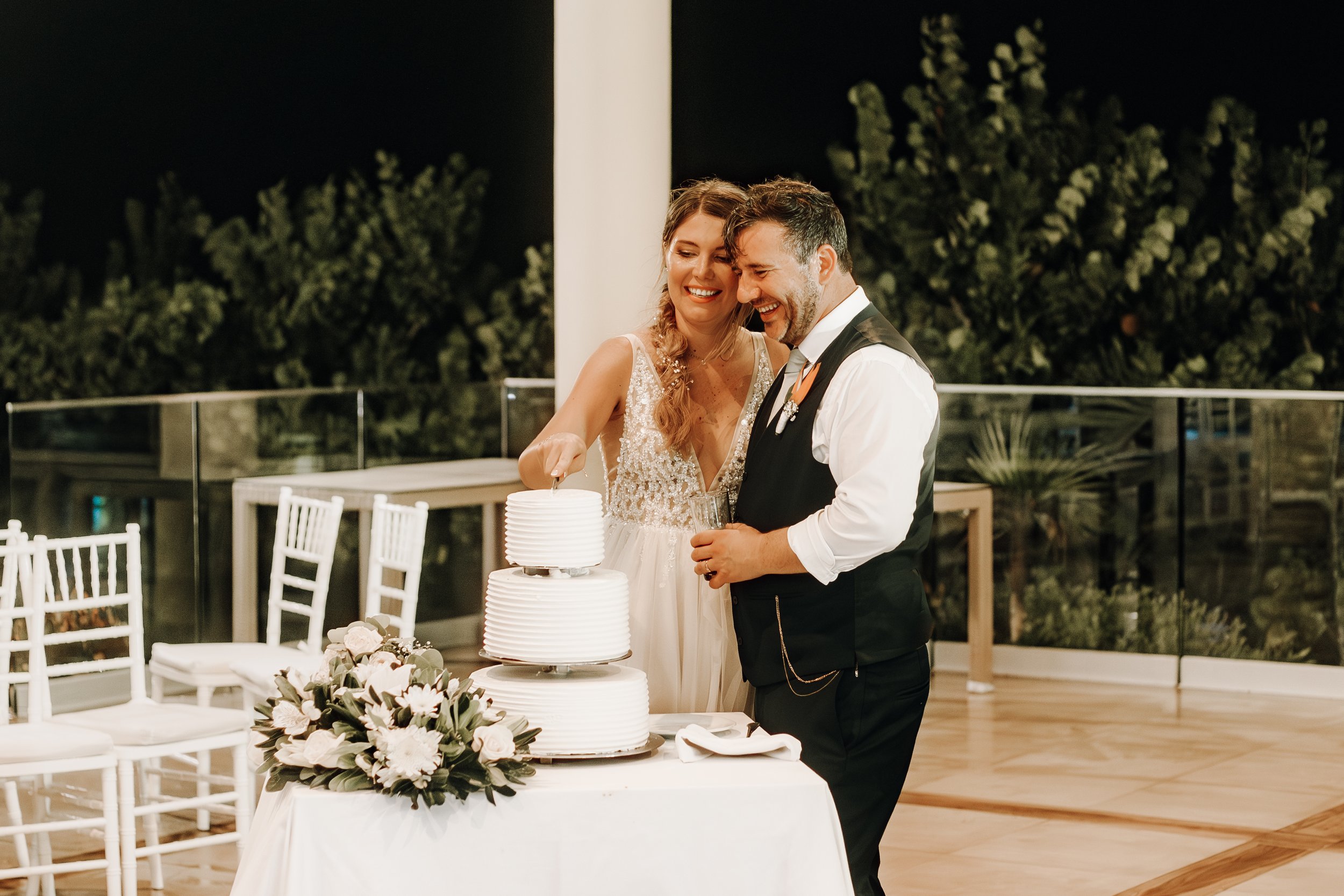 A newly married couple cutting their wedding cake, smiling, with a wedding bouquet on the table, during nighttime outdoor reception.