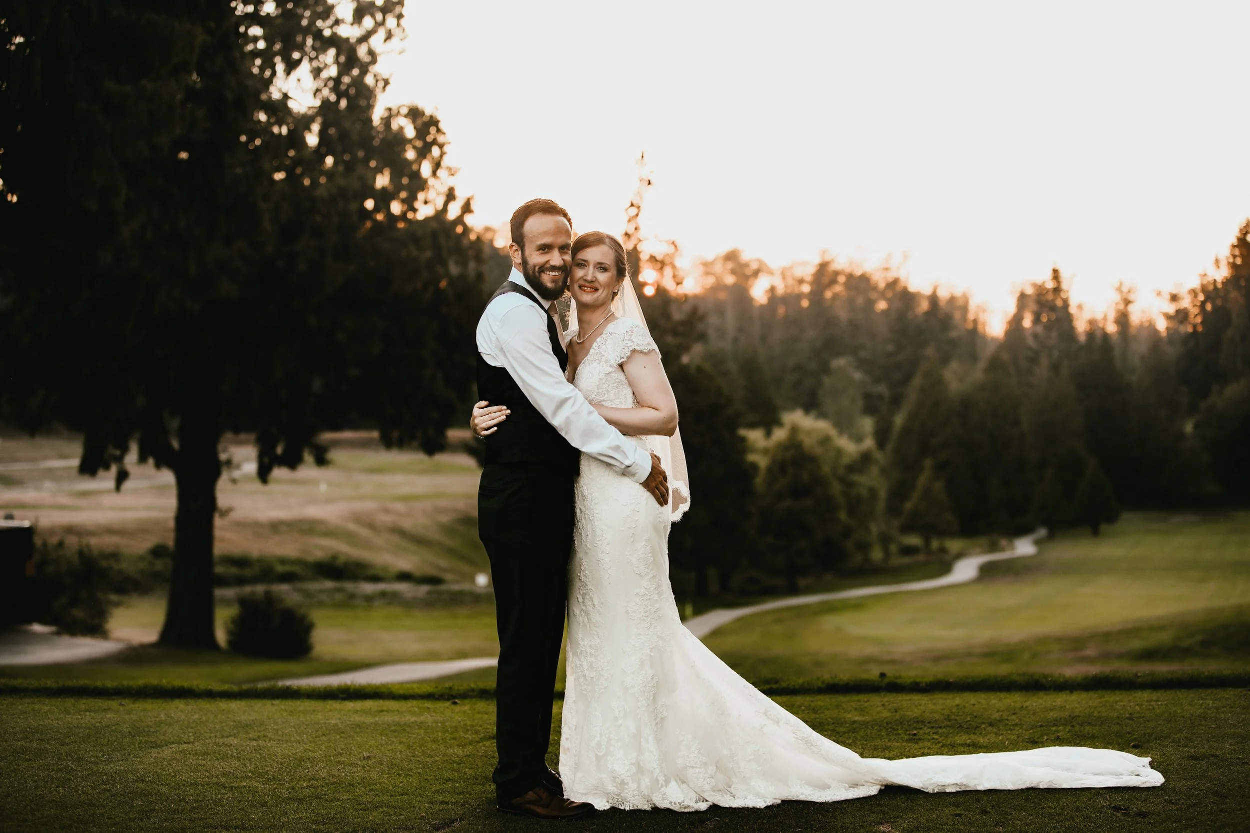 A newlywed couple standing on a lawn during sunset. The groom is wearing a black vest and pants with a white shirt, and the bride is in a lace wedding gown with a veil. They are smiling, embracing, and looking at the camera with a scenic park and tre