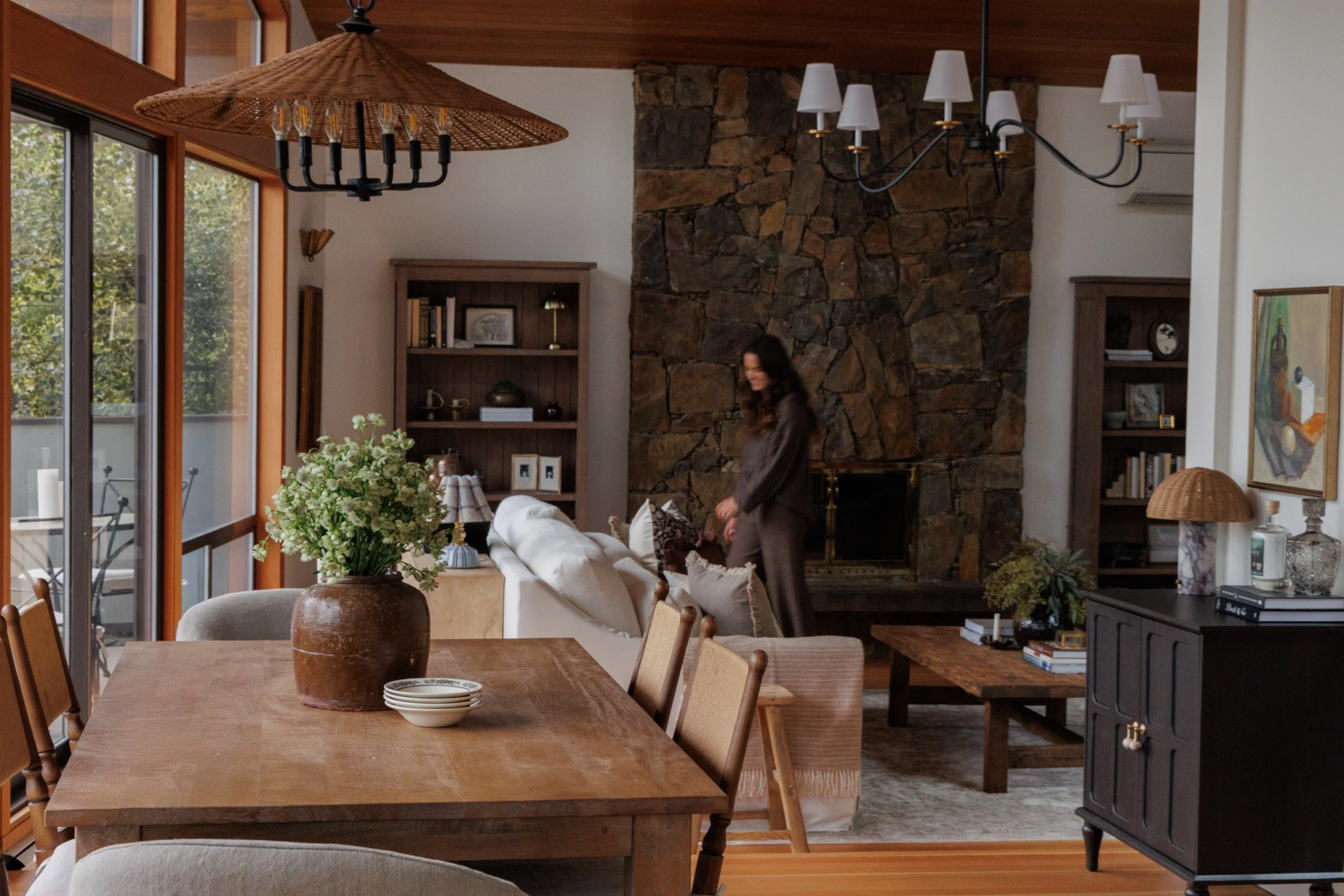 Living room with a wooden dining table, vases, a large flower arrangement, and a woman standing near the white sofa with a stone fireplace in the background.