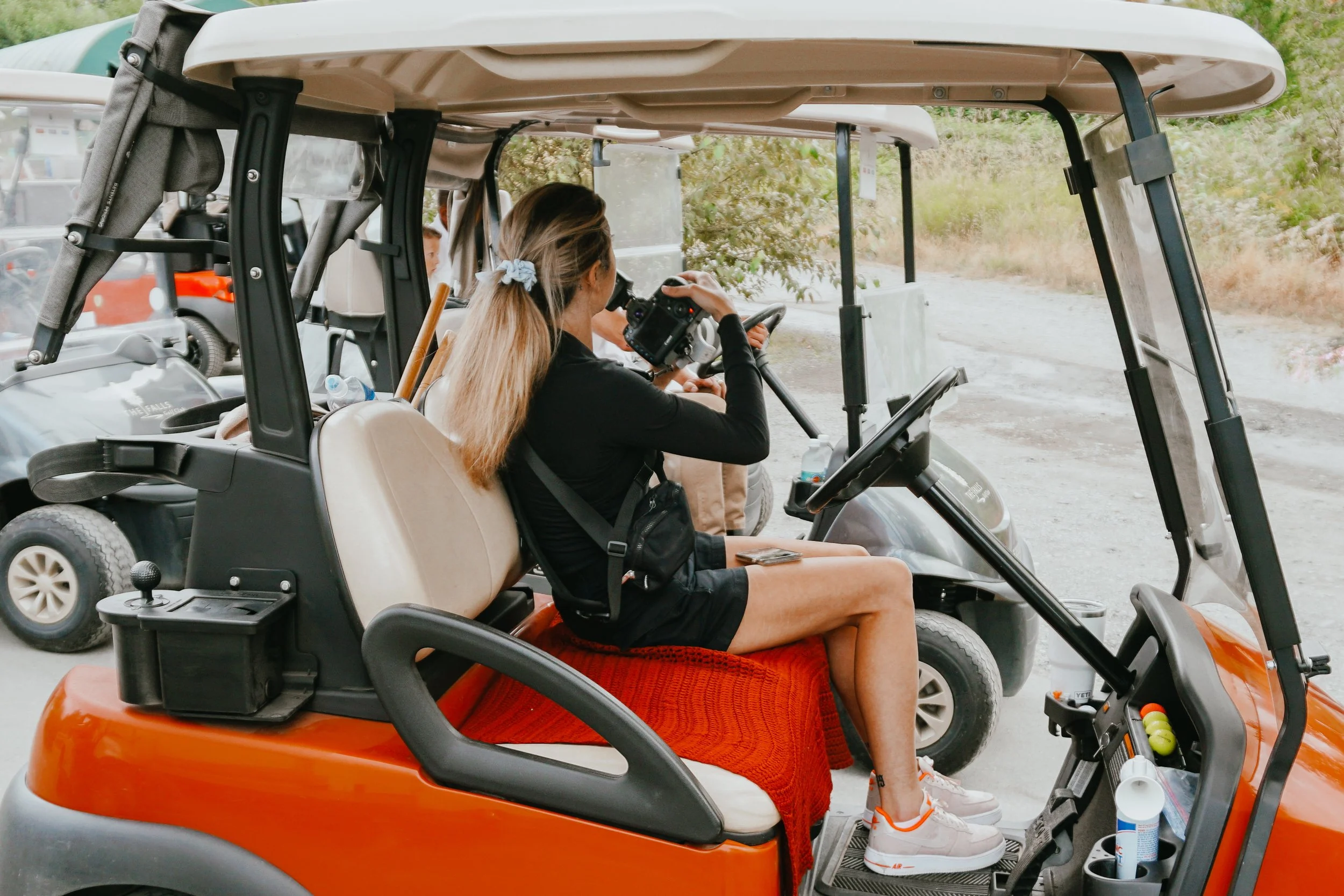 A woman with long hair in a ponytail, wearing a black shirt, shorts, and white sneakers, sitting in a golf cart, looking through a camera.