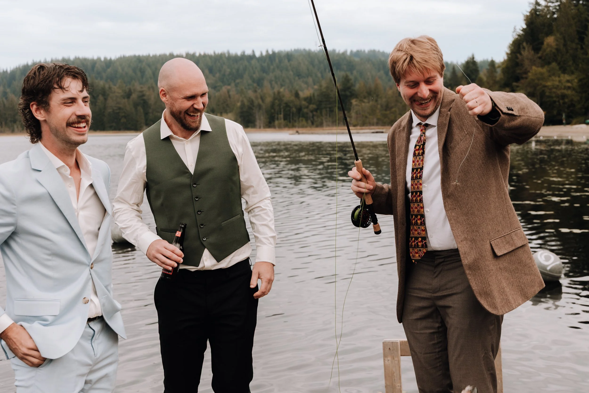 Three men dressed in formal attire, standing by a lake, with one of them fishing and smiling, the other two watching and laughing in the background, with trees and mountains under an overcast sky.