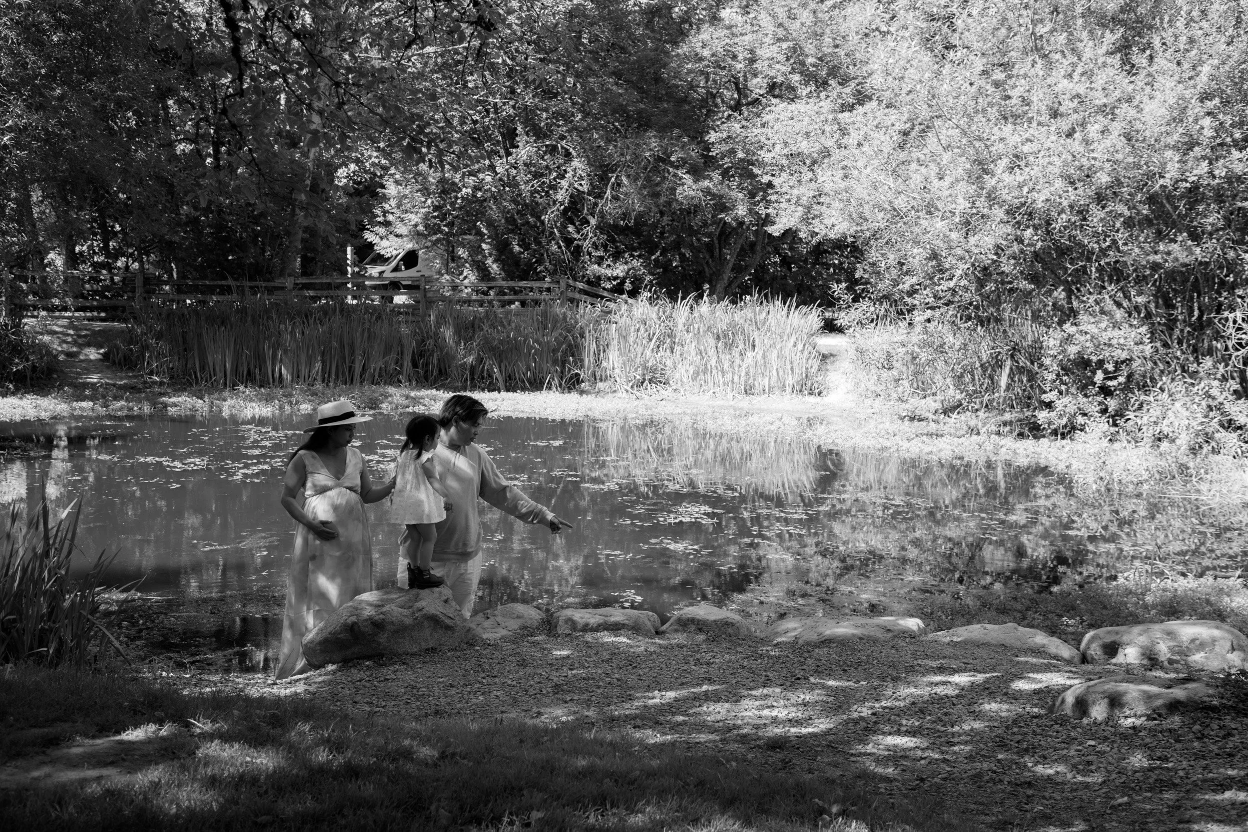 Three people, two women and a child, standing on rocks at the edge of a pond, with one woman and the child looking into the water and the other woman pointing towards it, surrounded by trees and tall grass.
