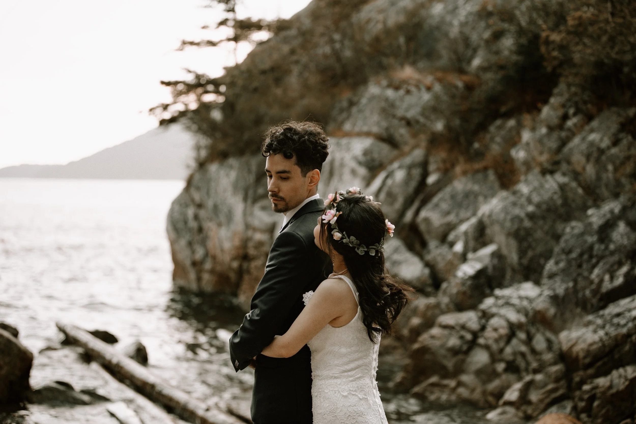 A bride and groom standing by the water on rocks with a rocky cliff in the background during sunset.