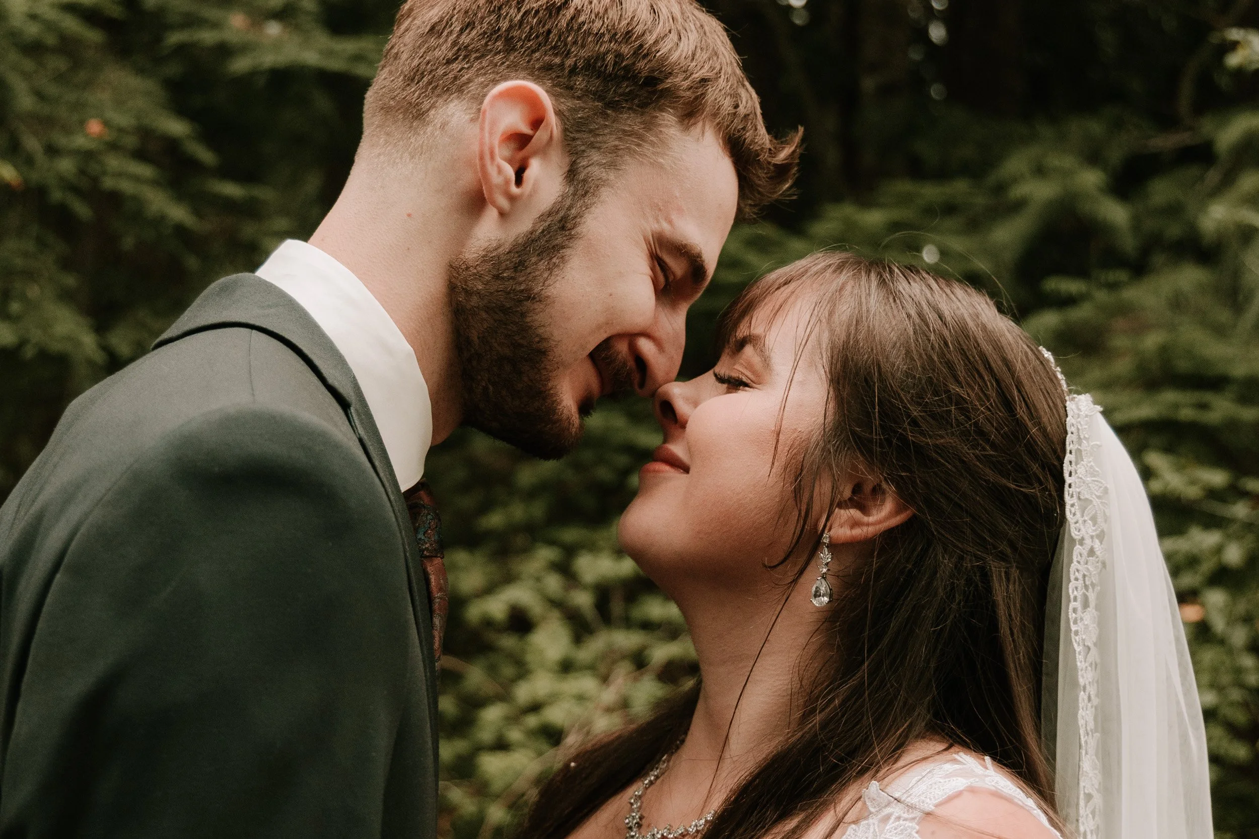 A bride and groom share an intimate moment on their wedding day, with their foreheads touching, eyes closed, surrounded by lush green foliage.