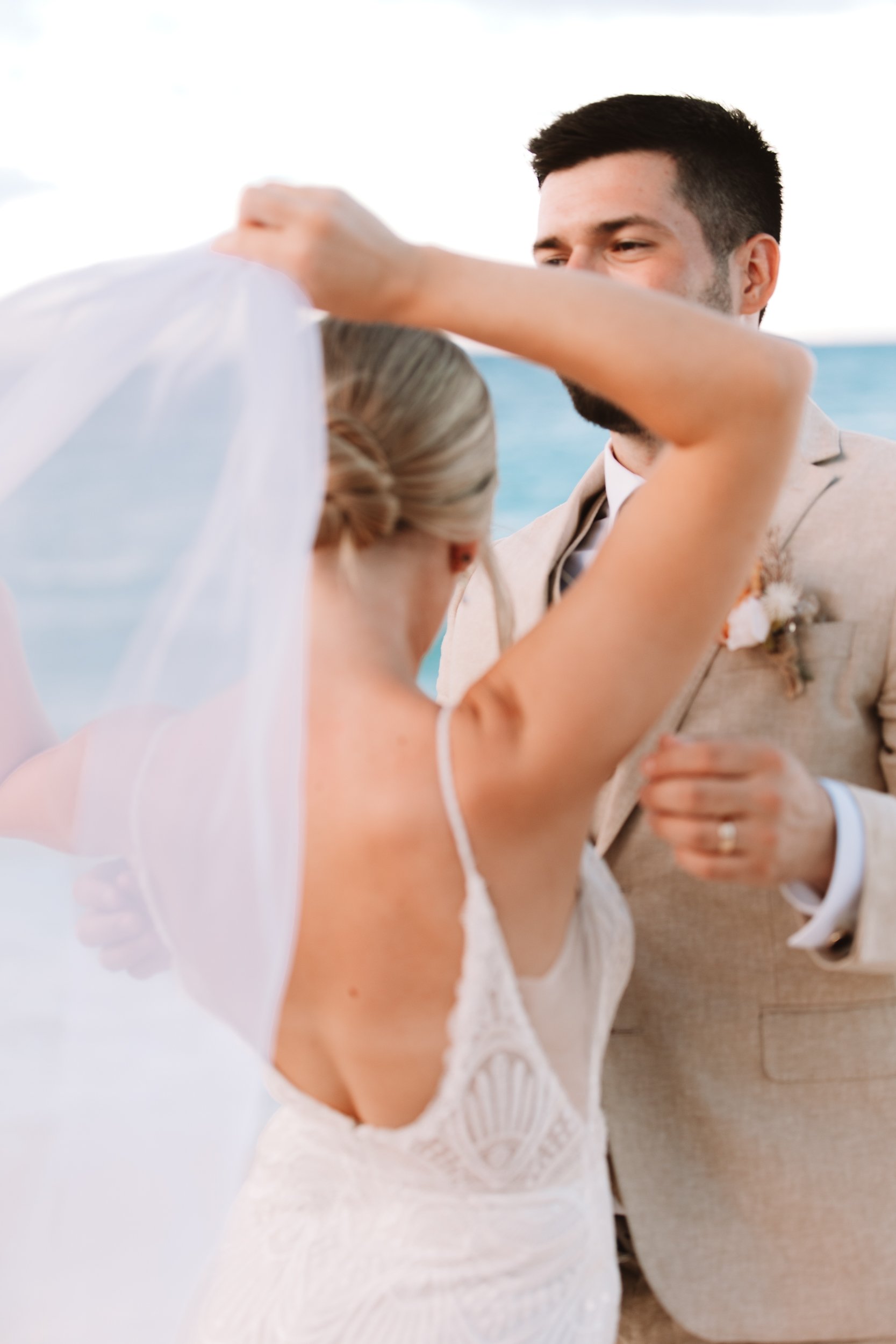 A bride and groom are standing on the beach, with the bride lifting her veil as the groom looks at her.