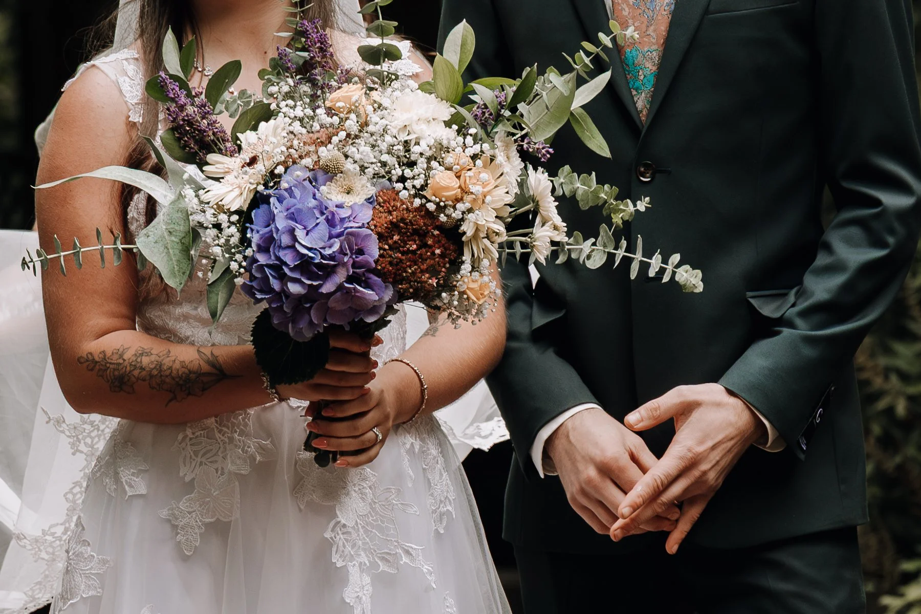Bride holding a bouquet of purple, white, and peach flowers, standing next to a groom in a dark suit with hands clasped.