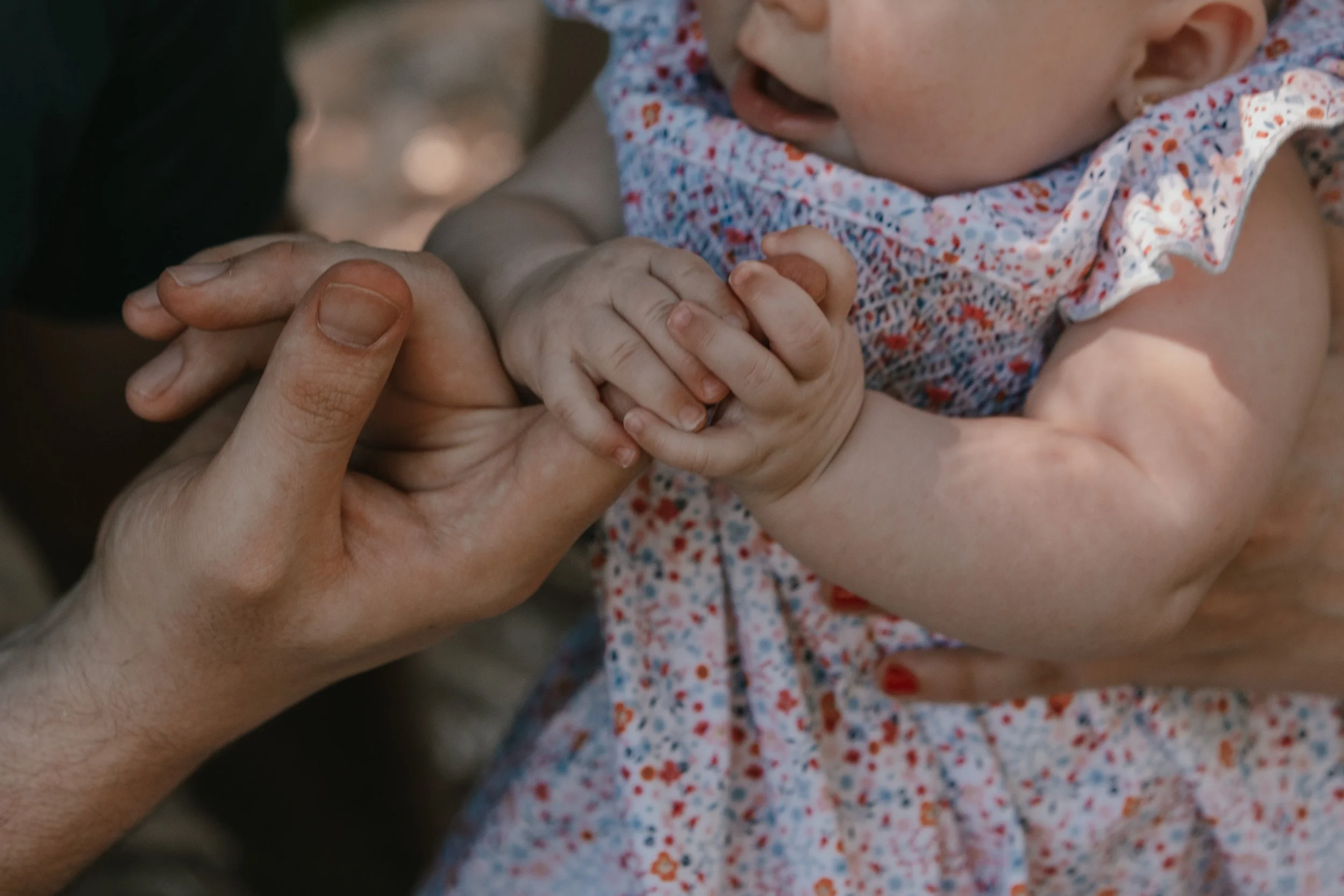 Close-up of an adult holding a baby's hands and arm, with the baby’s face partially visible, wearing a floral dress.