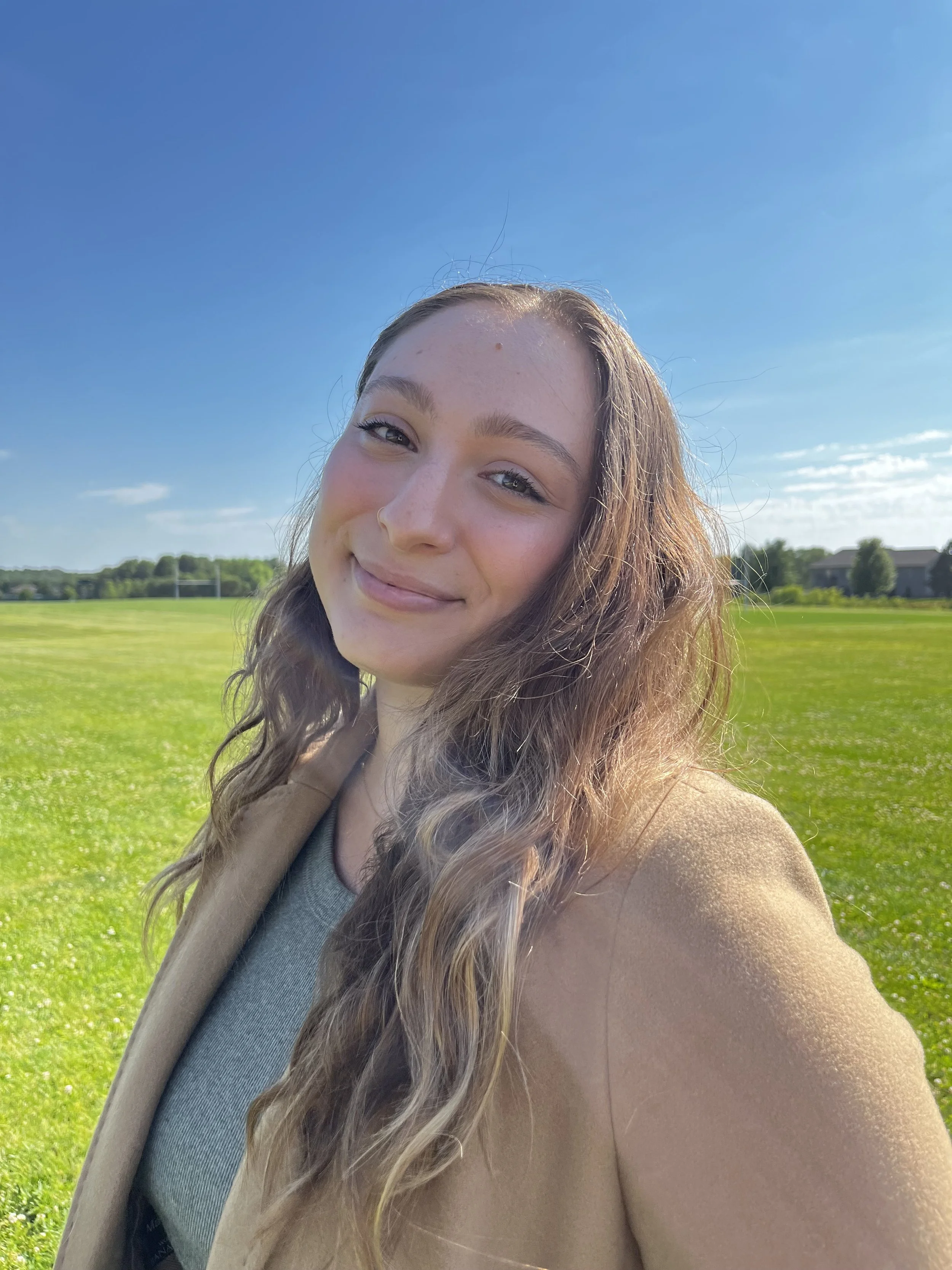 Young woman taking a selfie outdoors on a sunny day, standing on a grassy field with a blue sky and some buildings in the background.