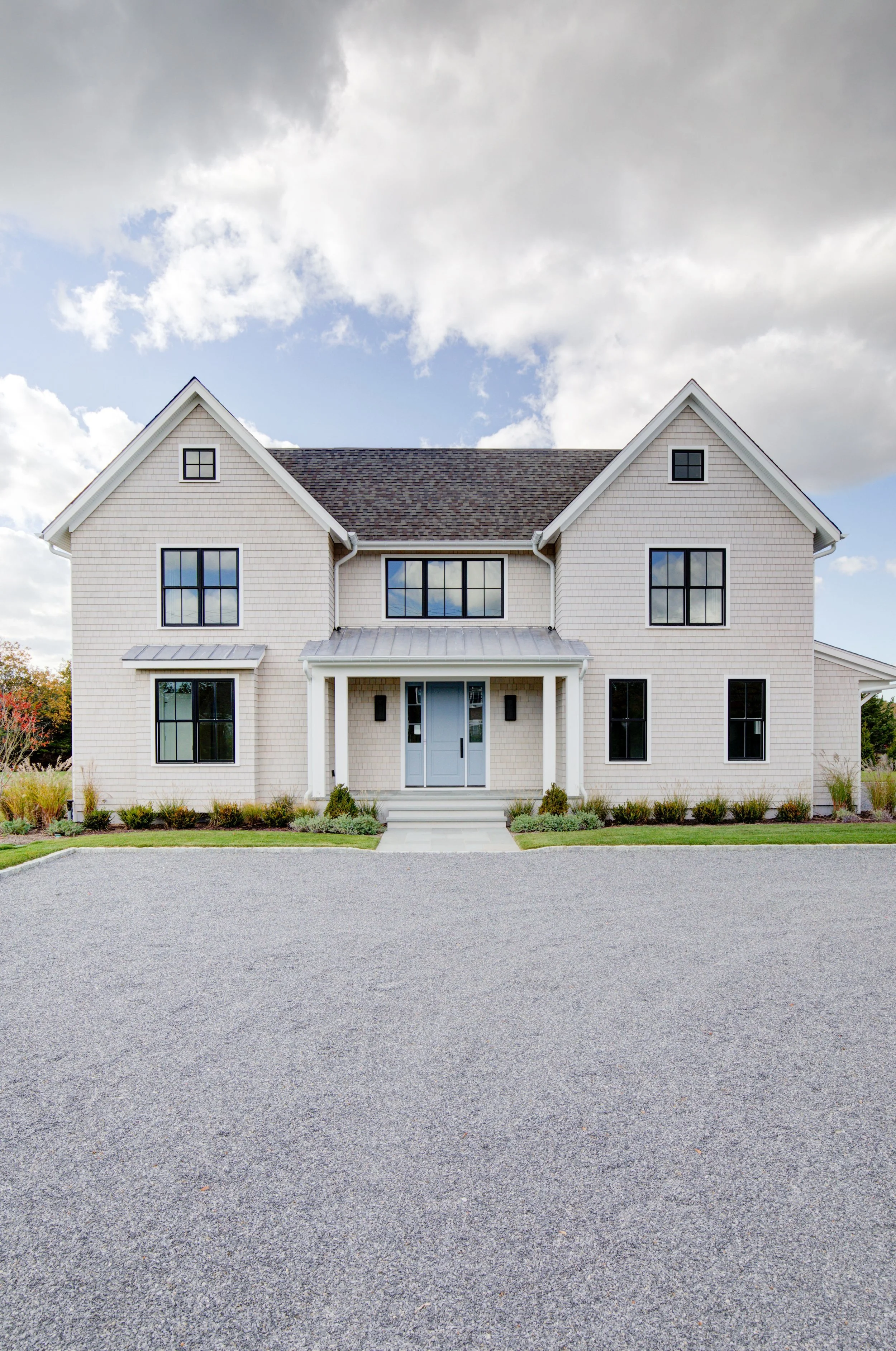 Modern two-story house with white exterior, black window frames, front porch, steps, and gravel driveway under cloudy sky.