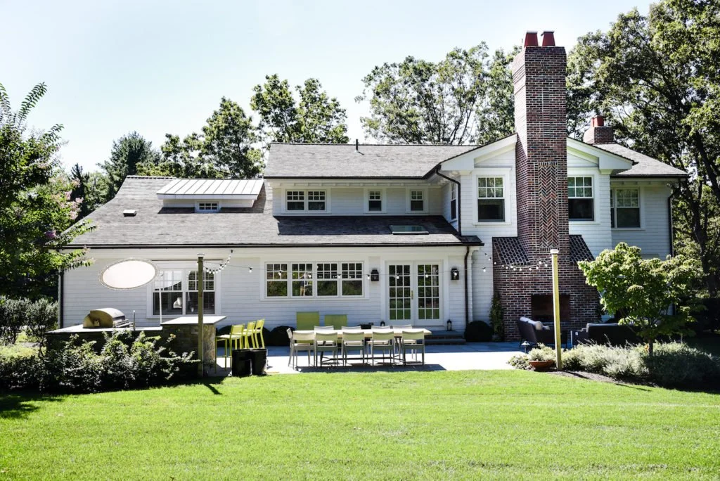 Backyard of a white two-story house with a large brick chimney, outdoor furniture, and a lush green lawn, surrounded by trees.