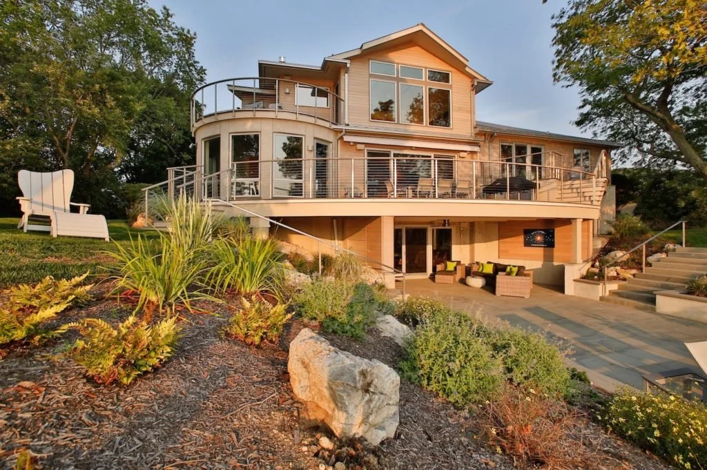 Large multi-story house with a wrap-around balcony and large glass windows, surrounded by landscaped garden with plants, rocks, and outdoor furniture.