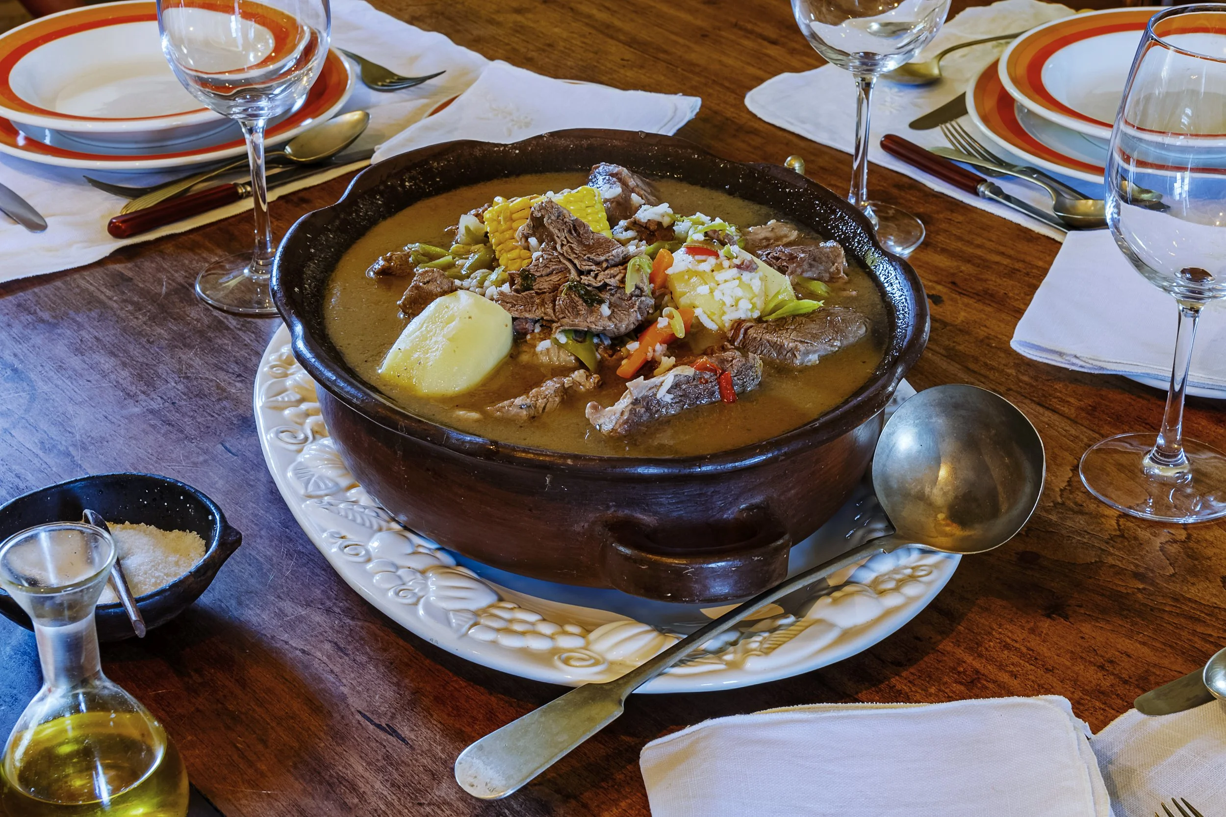 A large bowl of beef stew with vegetables, potatoes, and cornbread on a wooden dining table set for a meal.