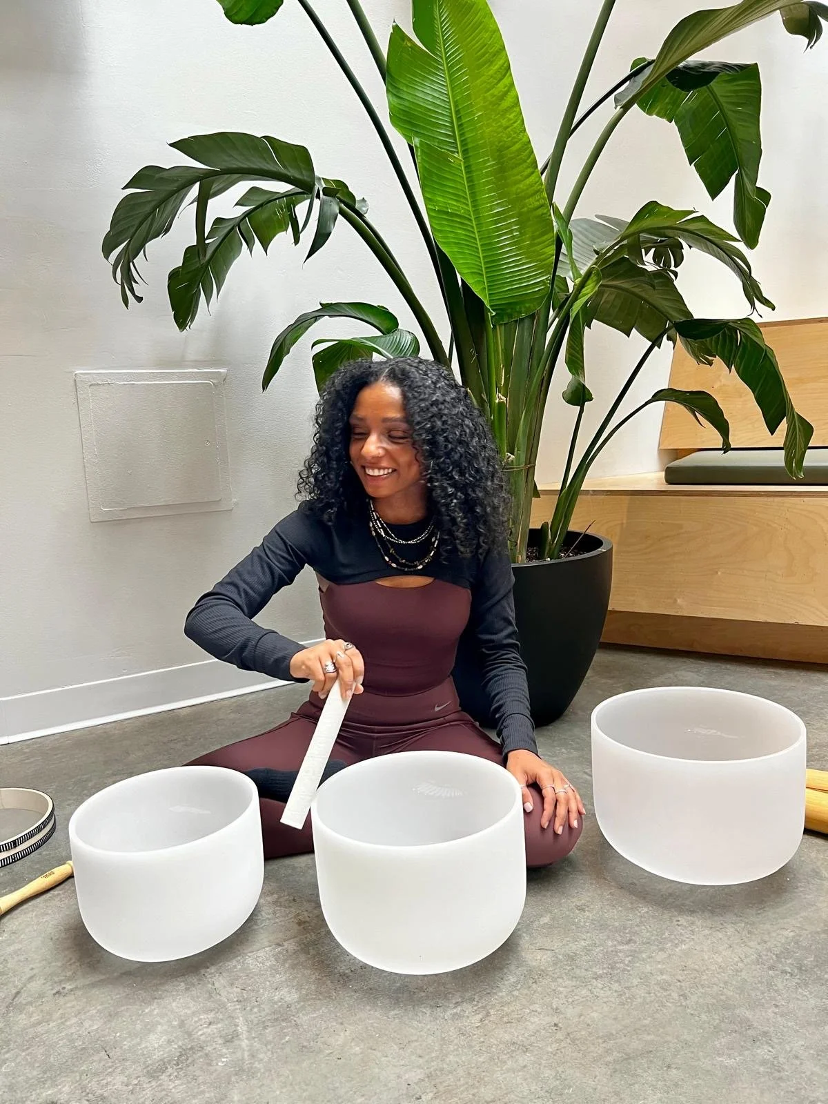 A woman with curly black hair sitting on the floor, smiling, playing crystal singing bowls, with a large green plant and a white wall in the background.