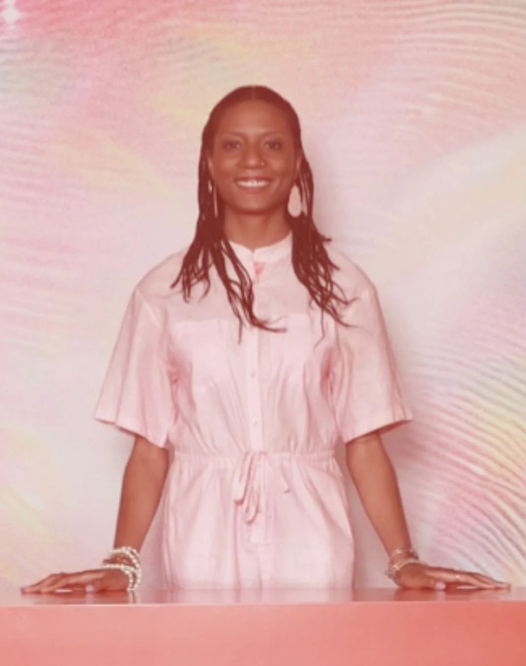 A smiling woman with long braided hair standing behind a pink table, wearing a pink dress with rolled-up sleeves and a drawstring waist, with a pastel swirl background.