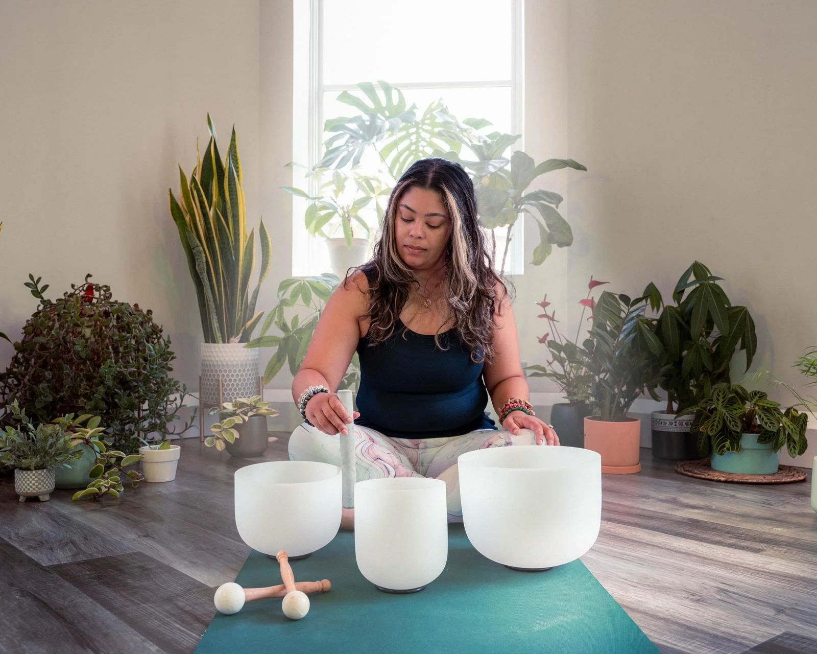 Woman playing crystal singing bowls in a room with potted plants, sitting on a yoga mat.
