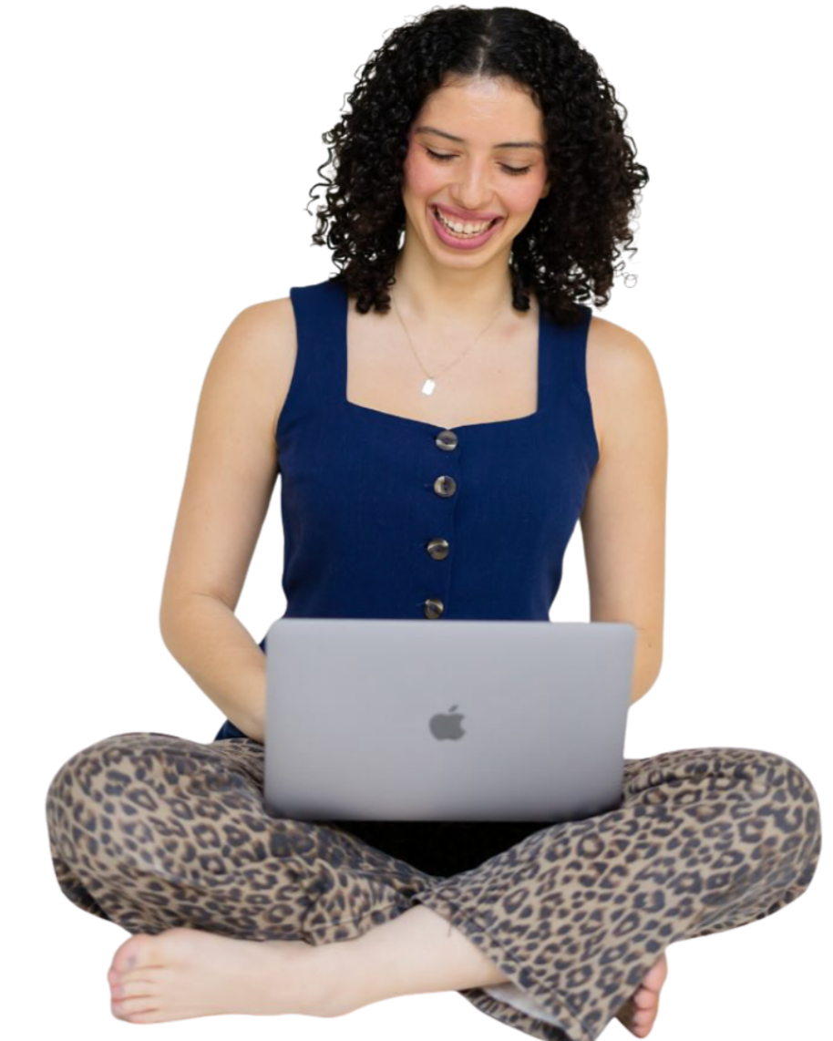A young woman with curly hair sitting cross-legged, smiling at her open MacBook laptop, wearing a blue sleeveless top and leopard print pants.