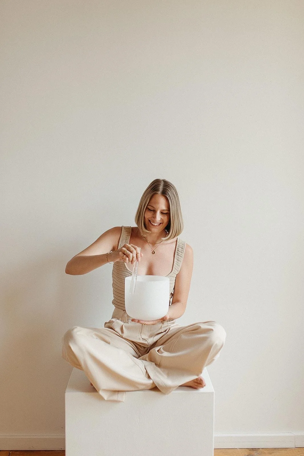 A woman sitting cross-legged on a white box, smiling and playing a crystal singing bowl.