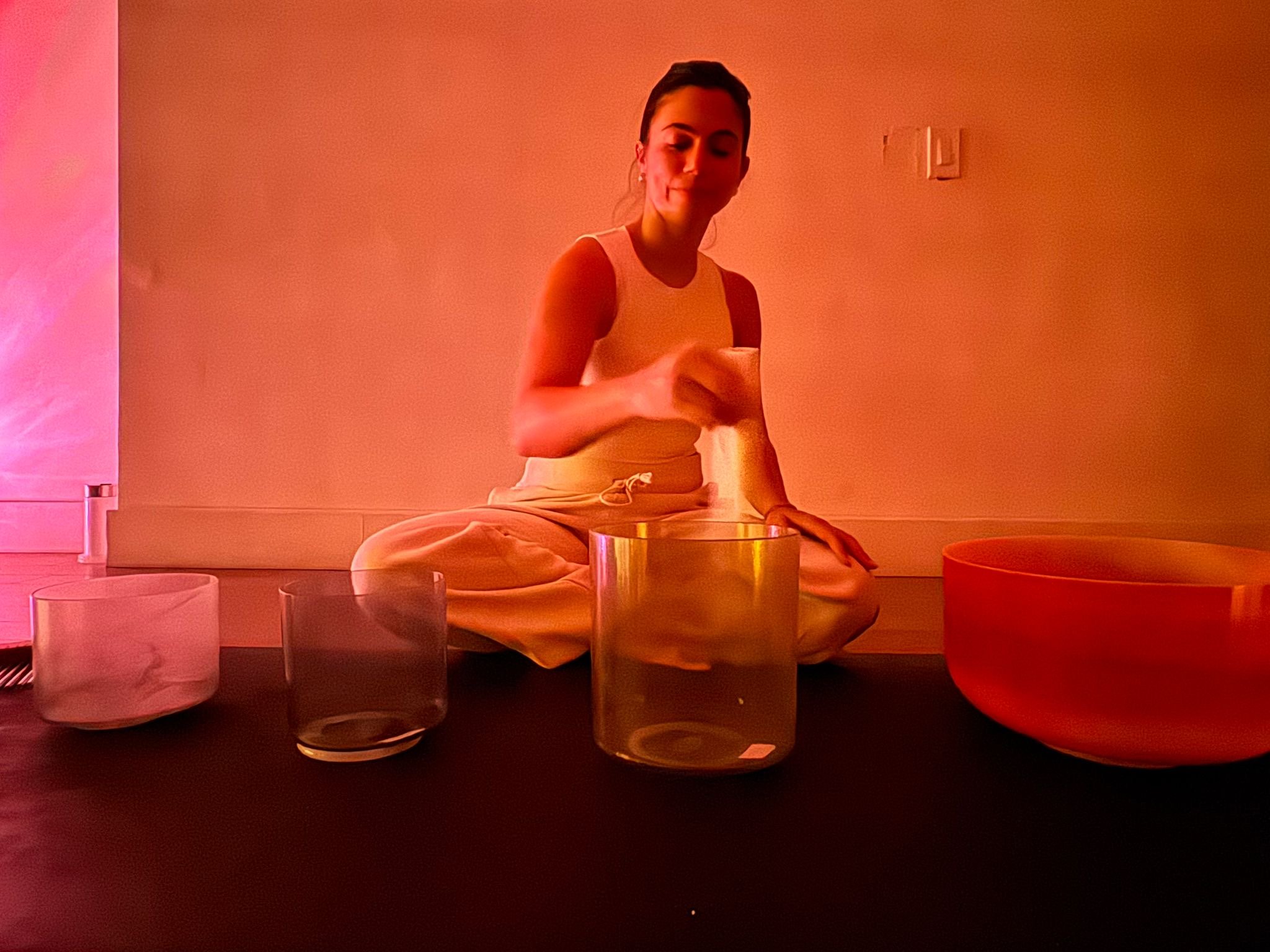 A woman sitting cross-legged on the floor, playing singing bowls of different sizes and colors in a dimly lit room.