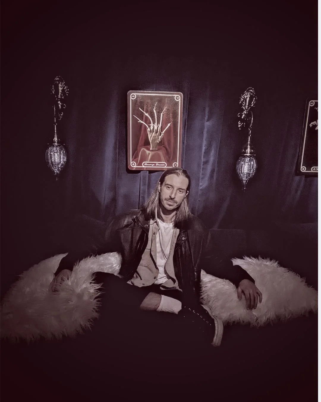 A man with long hair and a leather jacket sitting on a fluffy white rug in a dark, ornate room. Behind him are framed art pieces and decorative wall sconces.
