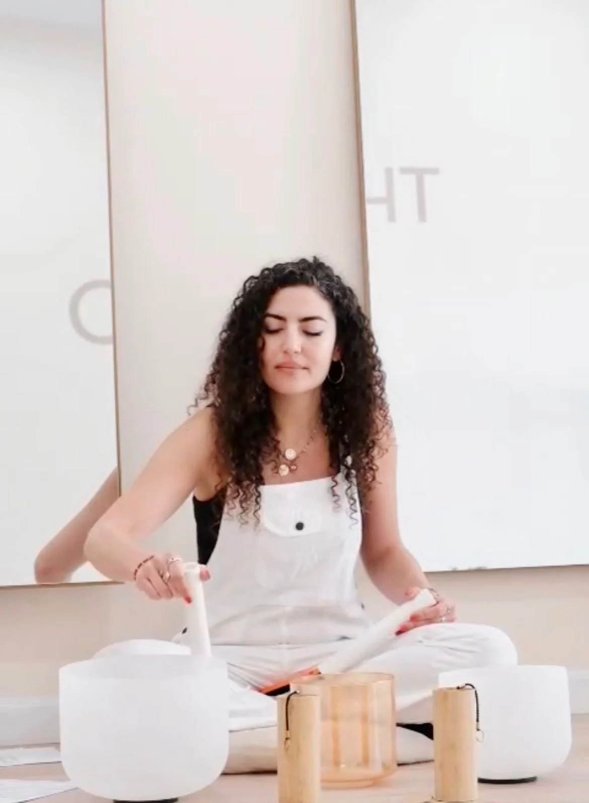 A young woman with curly hair and hoop earrings is sitting cross-legged on the floor, playing singing bowls with a mallet.