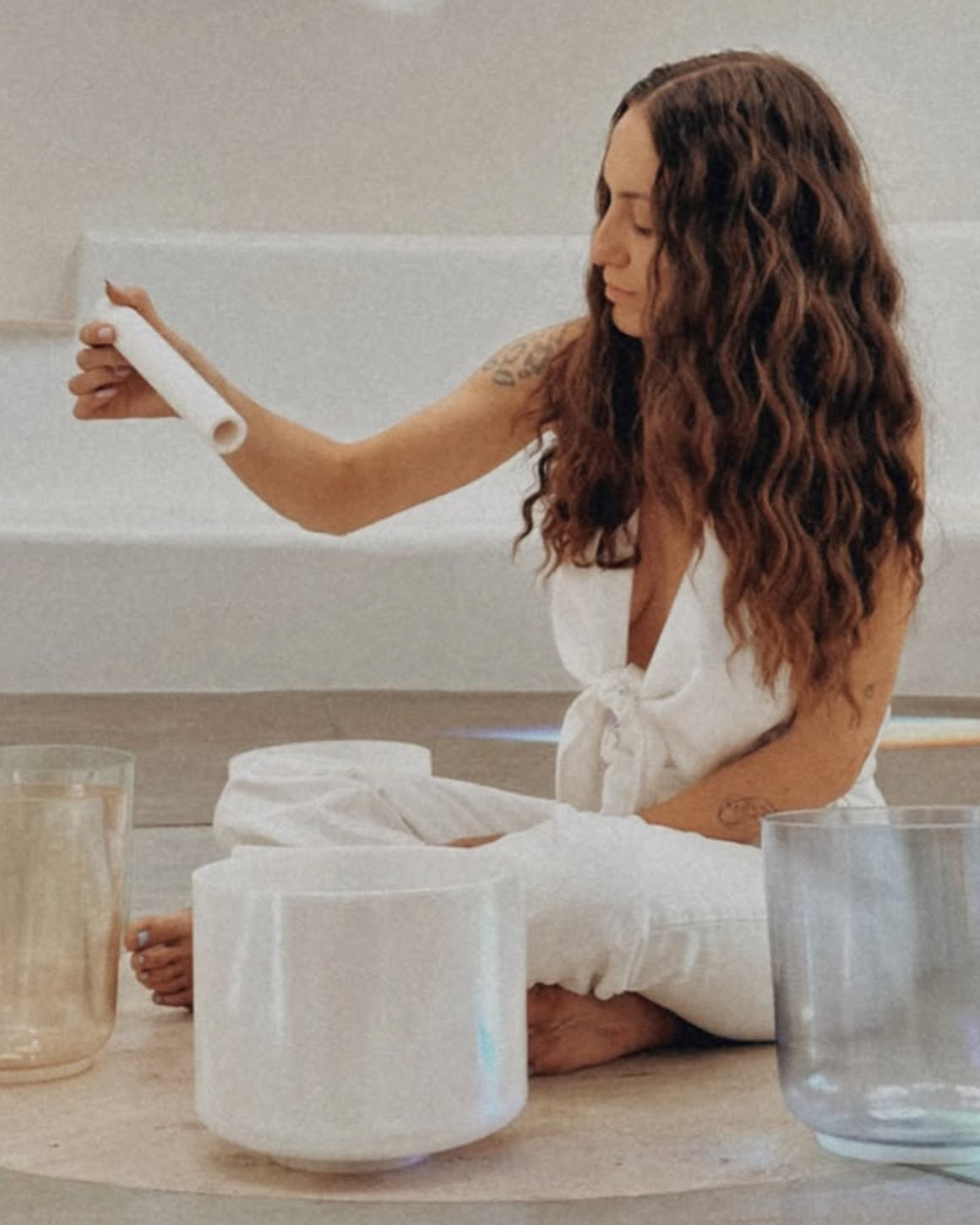 A woman with long wavy hair sitting cross-legged on the floor, holding a white candle, surrounded by several large candles in various colors.