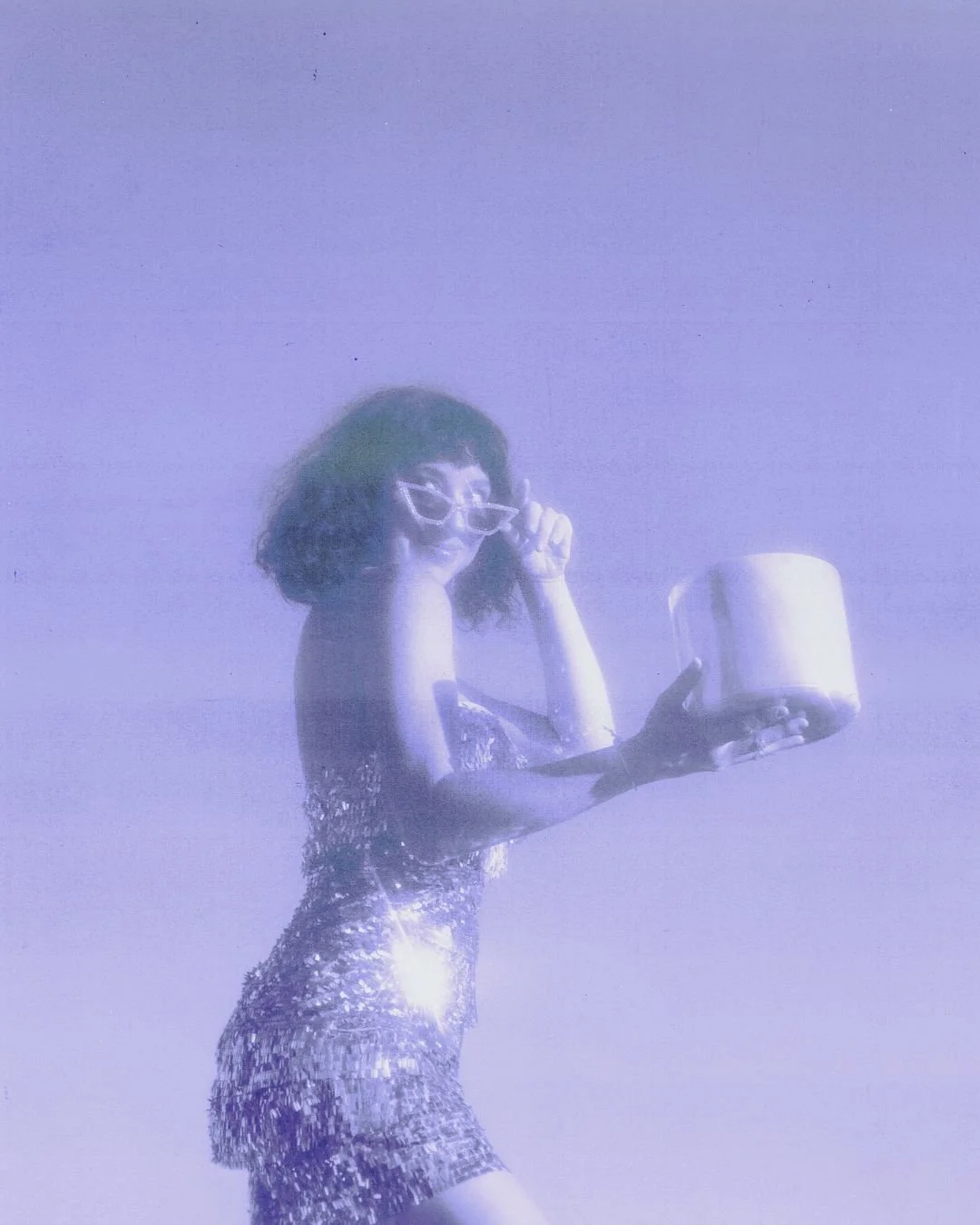 A woman with curly hair and glasses in a shiny, sparkly dress holding a container, against a plain background.