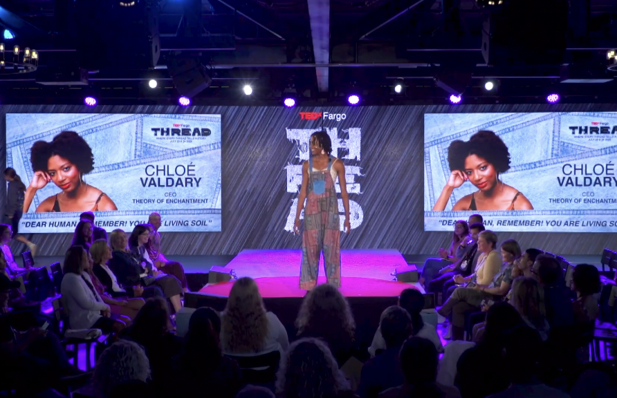A woman standing on stage at a TEDx event speaking to an audience. Large screens behind her display her name, title, and a photo, with the name "Chloé Valdary" and the text "CEO, Theory of Enchantment." The stage has purple and pink lighting, with an audience seated on both sides.