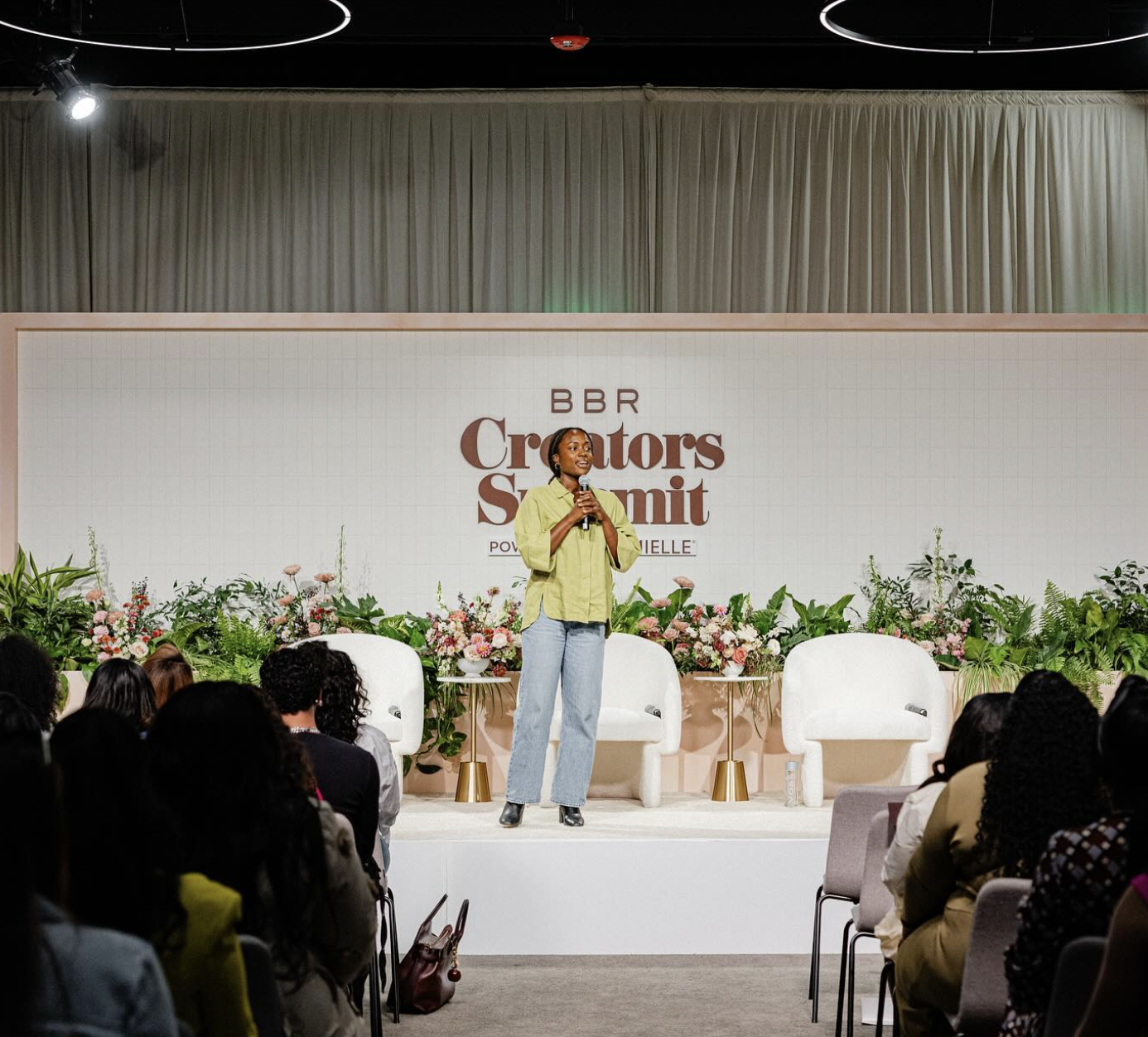 A woman standing on stage speaking into a microphone at the BBR Creators Summit, with an audience seated in front of her, and a backdrop decorated with flowers and the event logo.