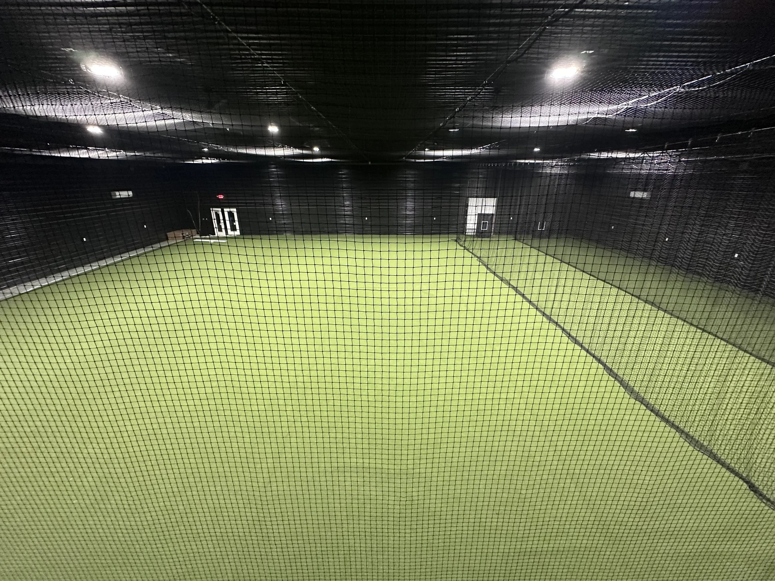 Indoor batting cage with artificial turf and black netting overhead.