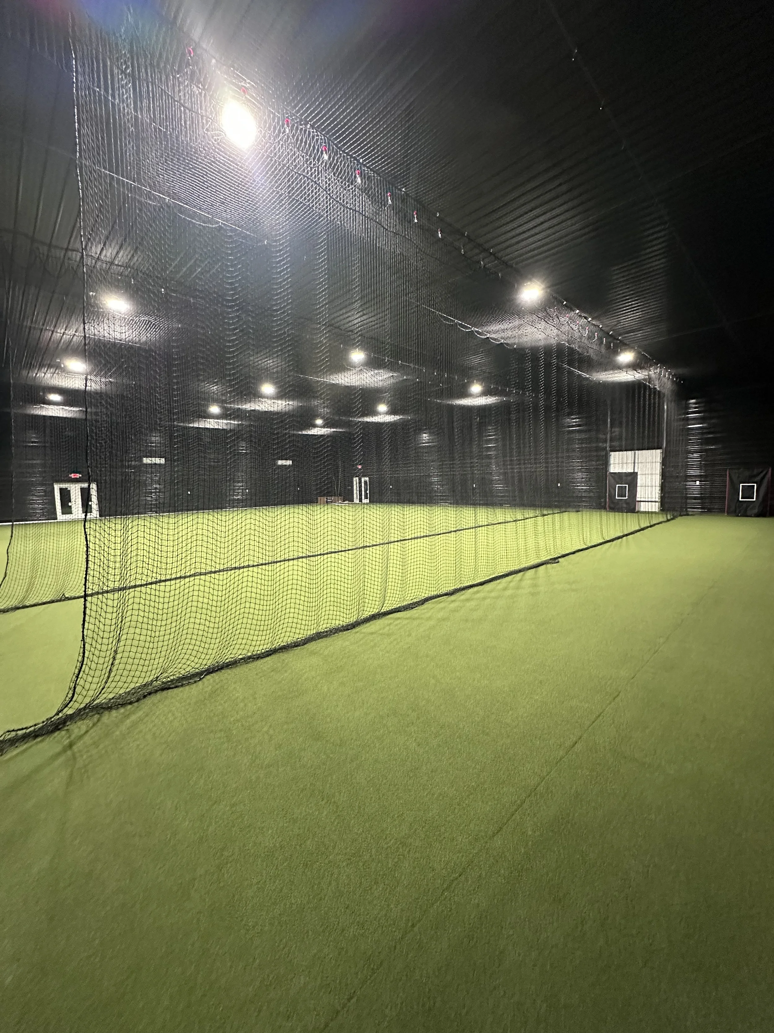 Indoor sports facility with artificial turf, sport netting, and nilluminated by overhead lights.