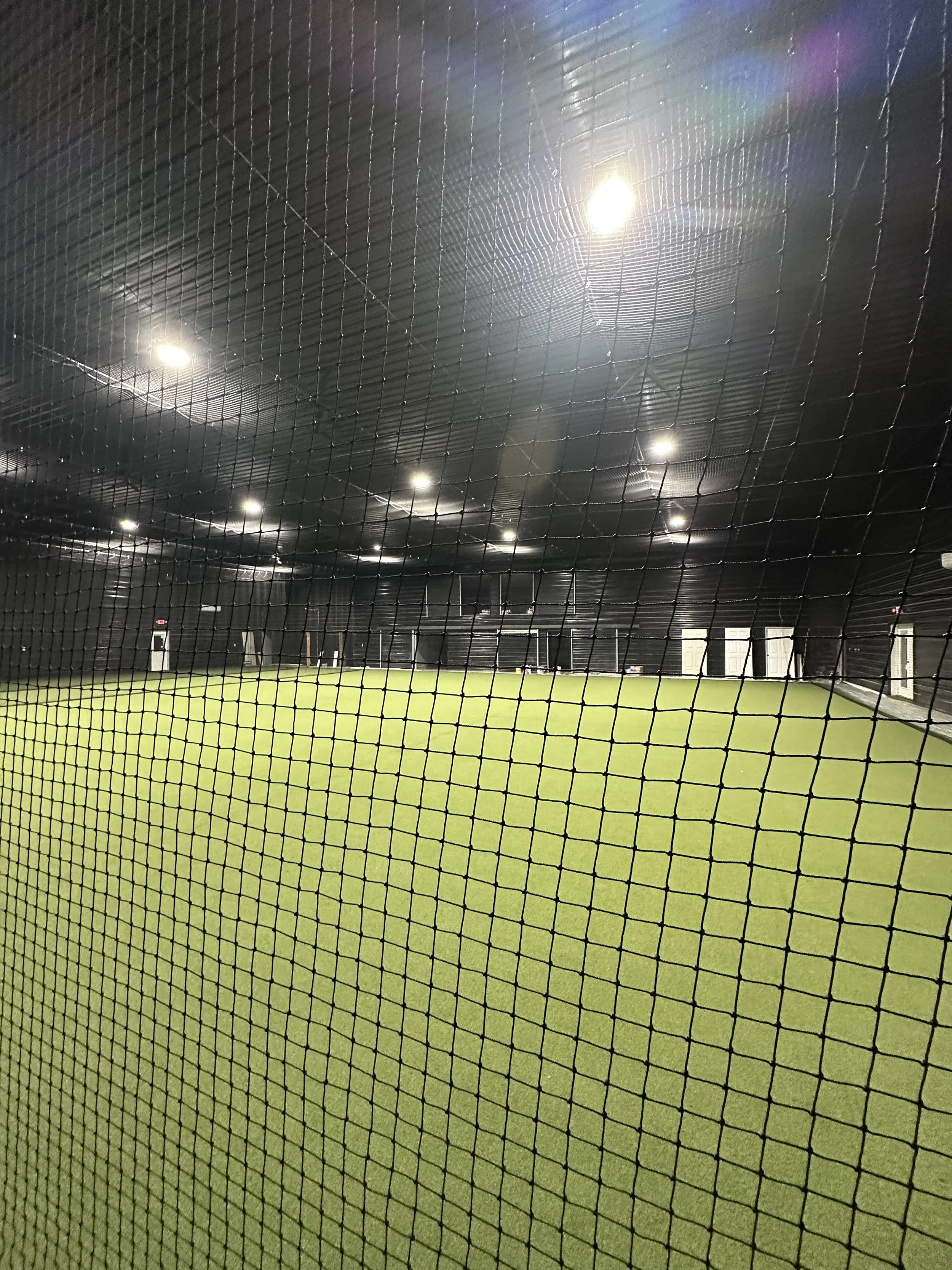 A view of an indoor sports facility with artificial turf, tall ceiling, and bright ceiling lights seen through a black sport netting.