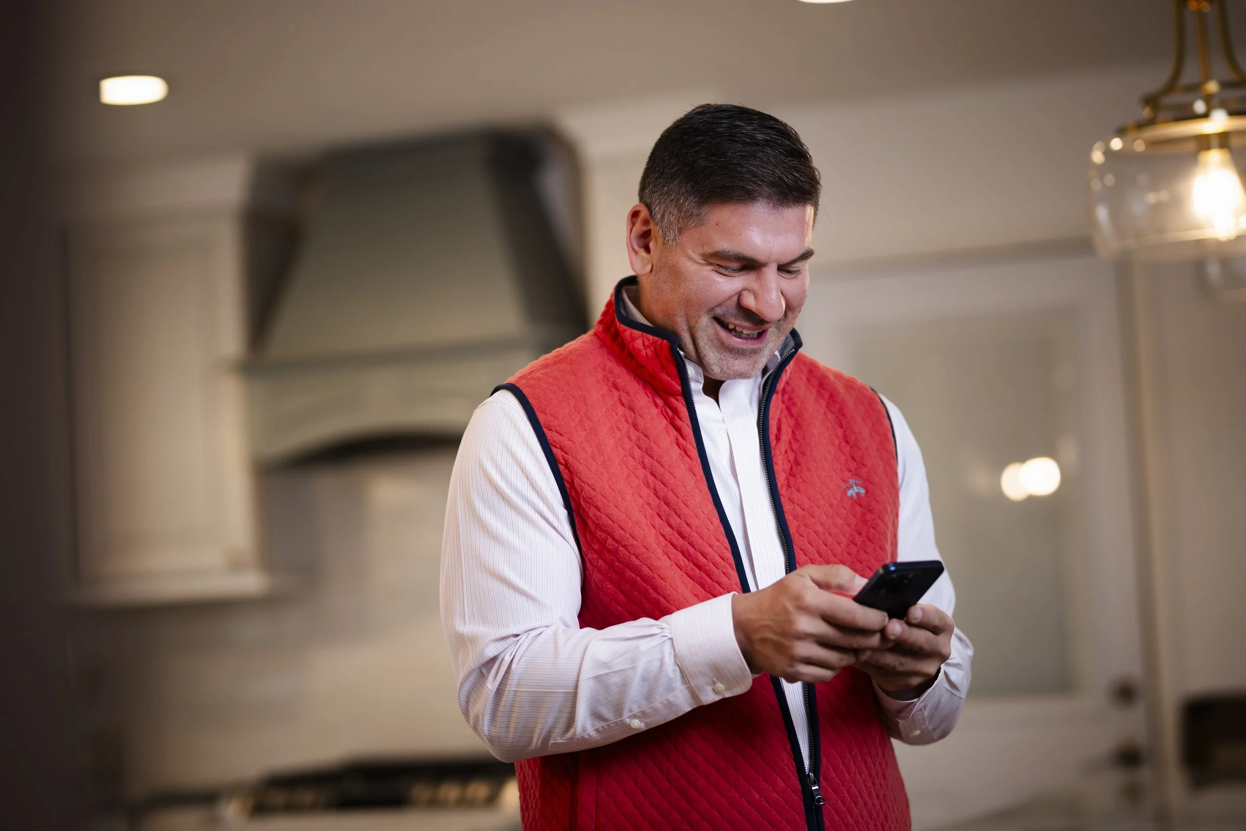 A man smiling while looking at his phone in a kitchen.