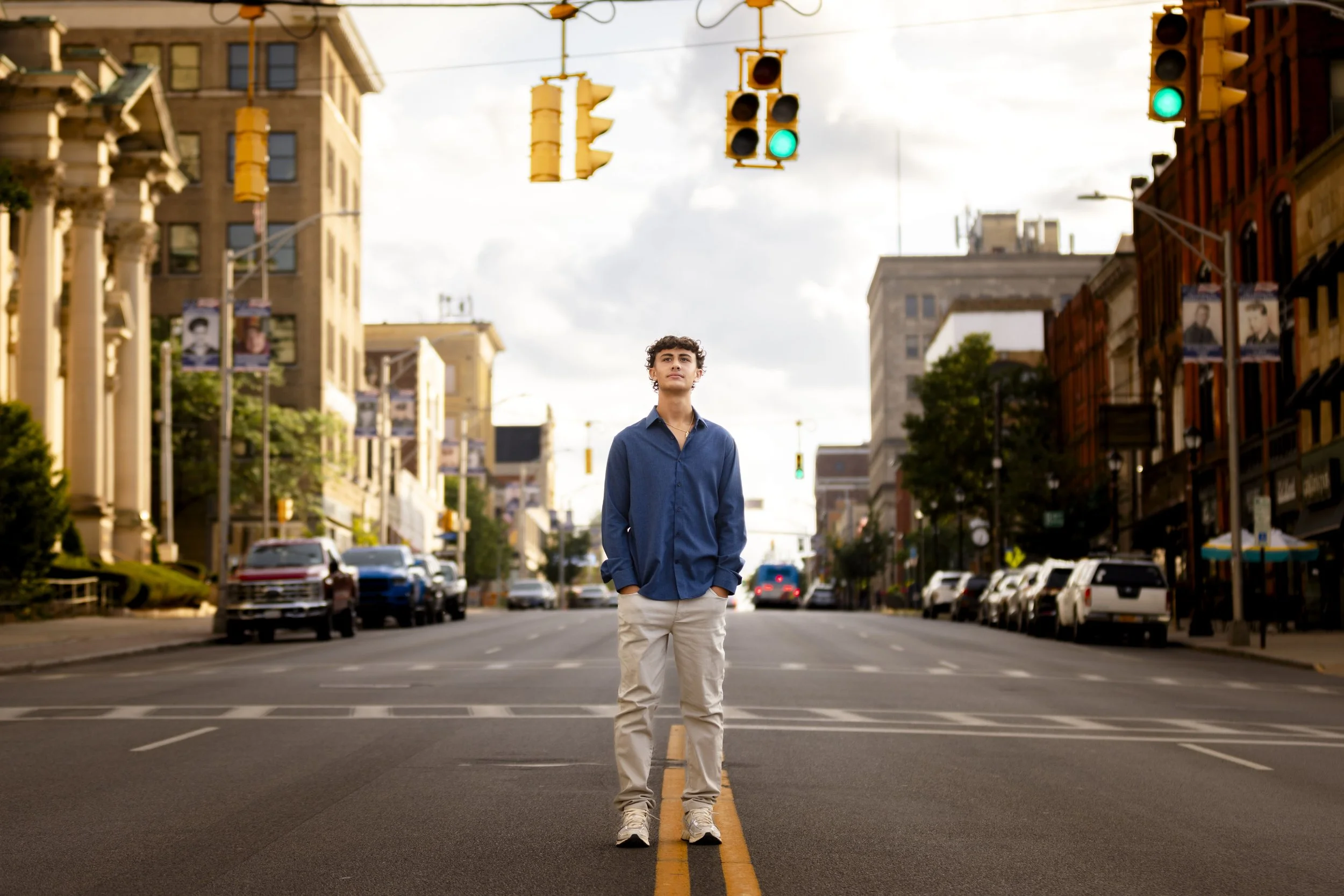 A young man standing in the middle of an empty city street with his hands in his pockets, looking up. The street is surrounded by buildings and parked cars, with traffic lights overhead showing green.