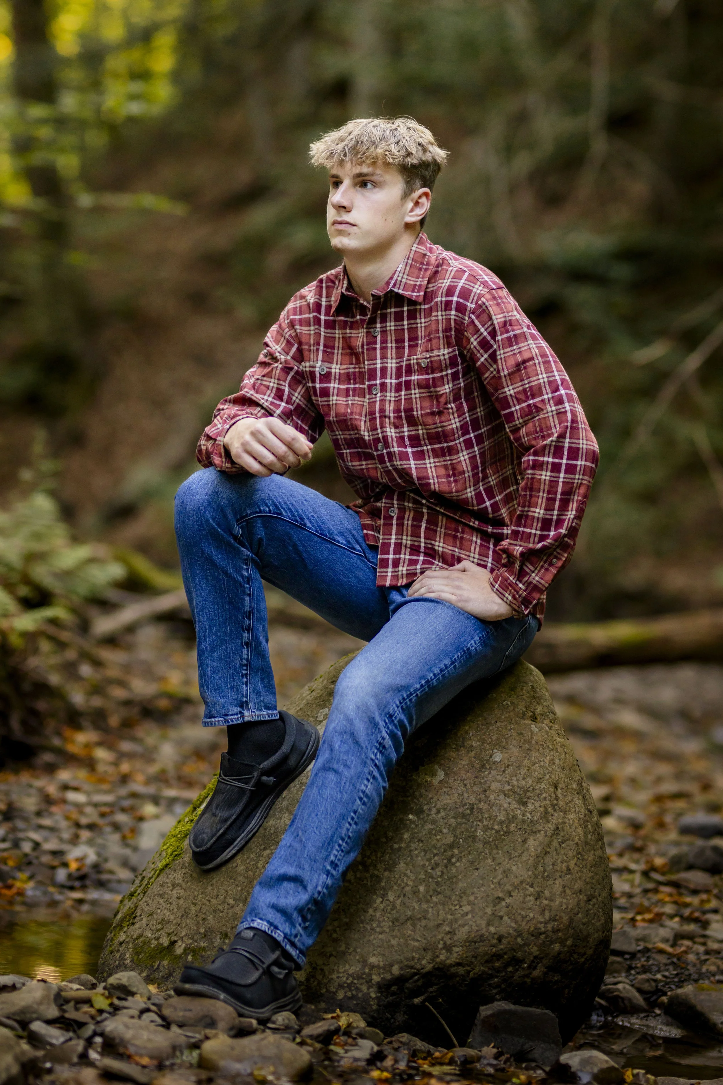 A young man wearing a red plaid shirt, blue jeans, and black shoes sitting on a large moss-covered rock in a forest setting.