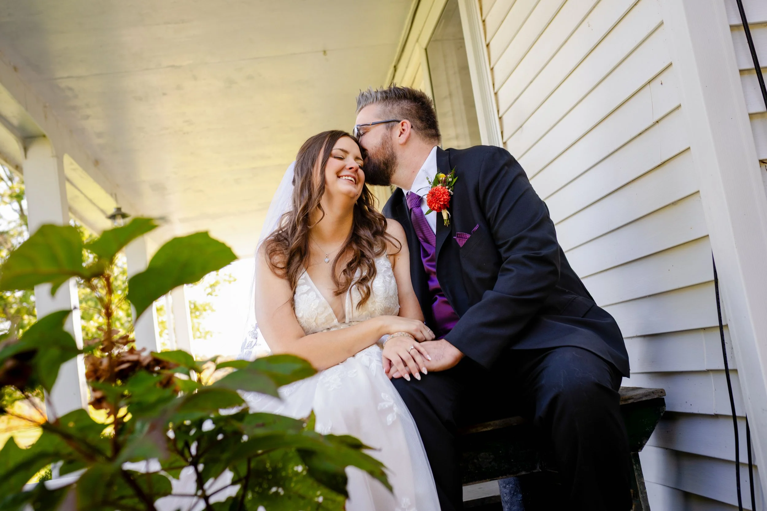 A groom and bride share a kiss on a porch, sitting on a bench, with greenery in the foreground.