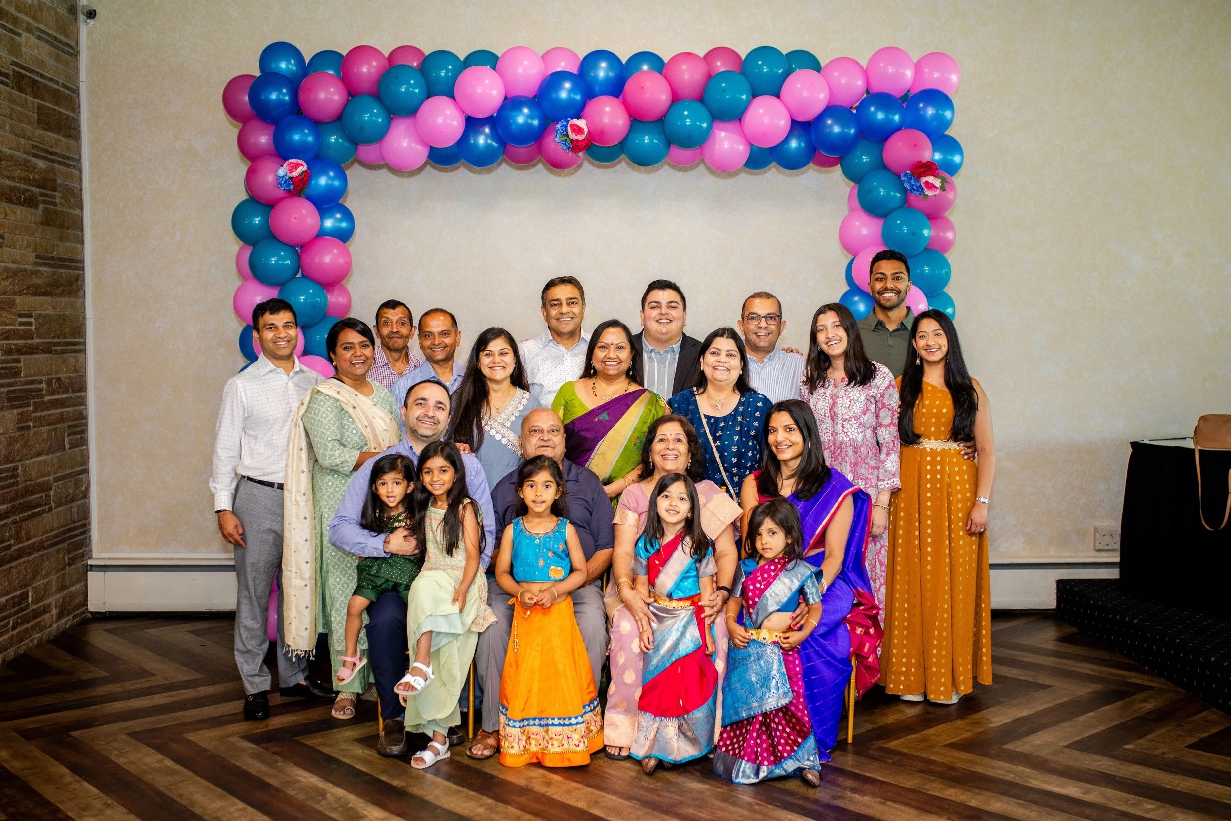 Group of adults and children posing at a celebration, standing in front of a colorful balloon arch with pink, blue, and purple balloons.