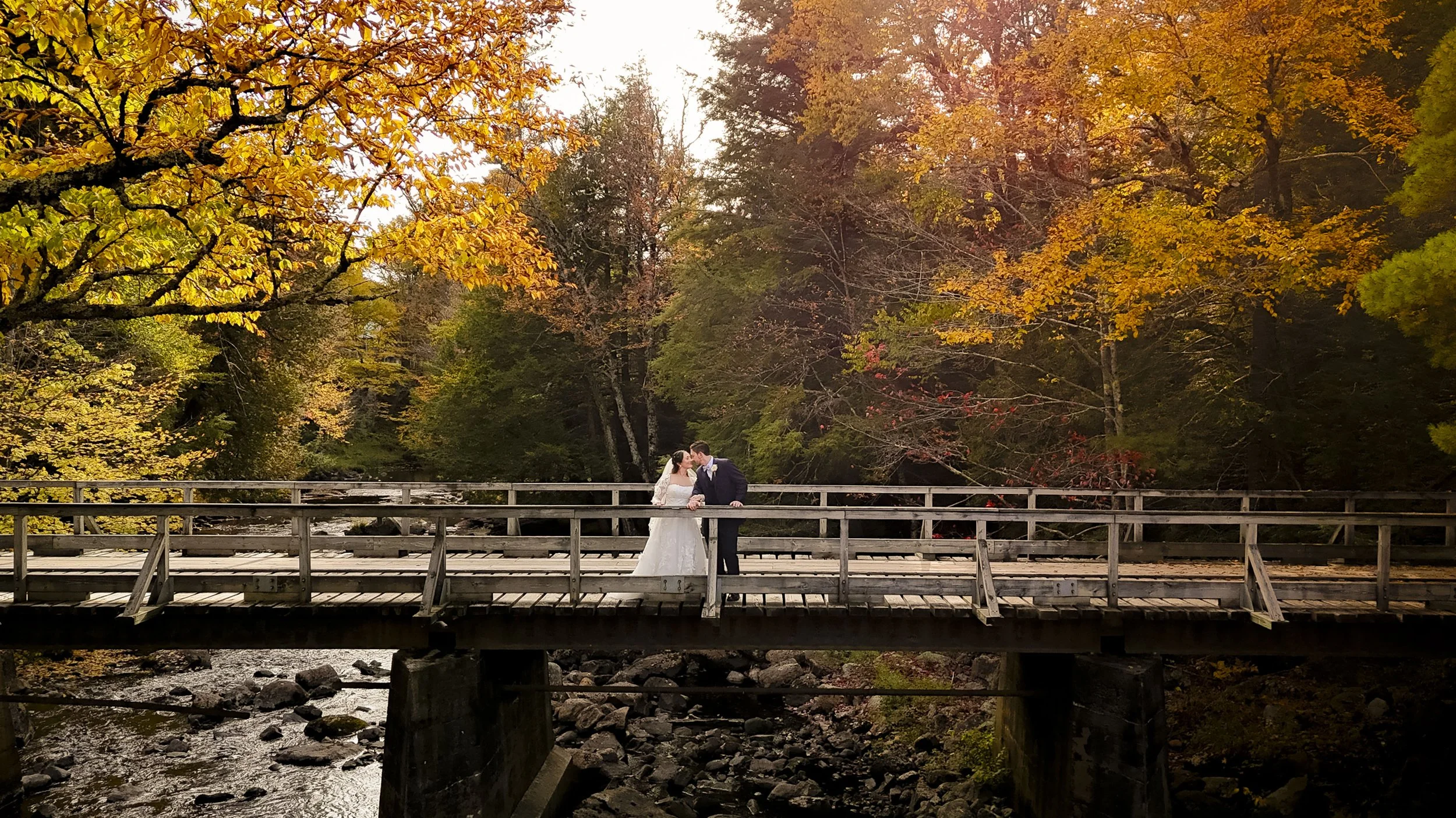 A bride and groom standing on a wooden bridge during fall, surrounded by colorful autumn trees, sharing a kiss.