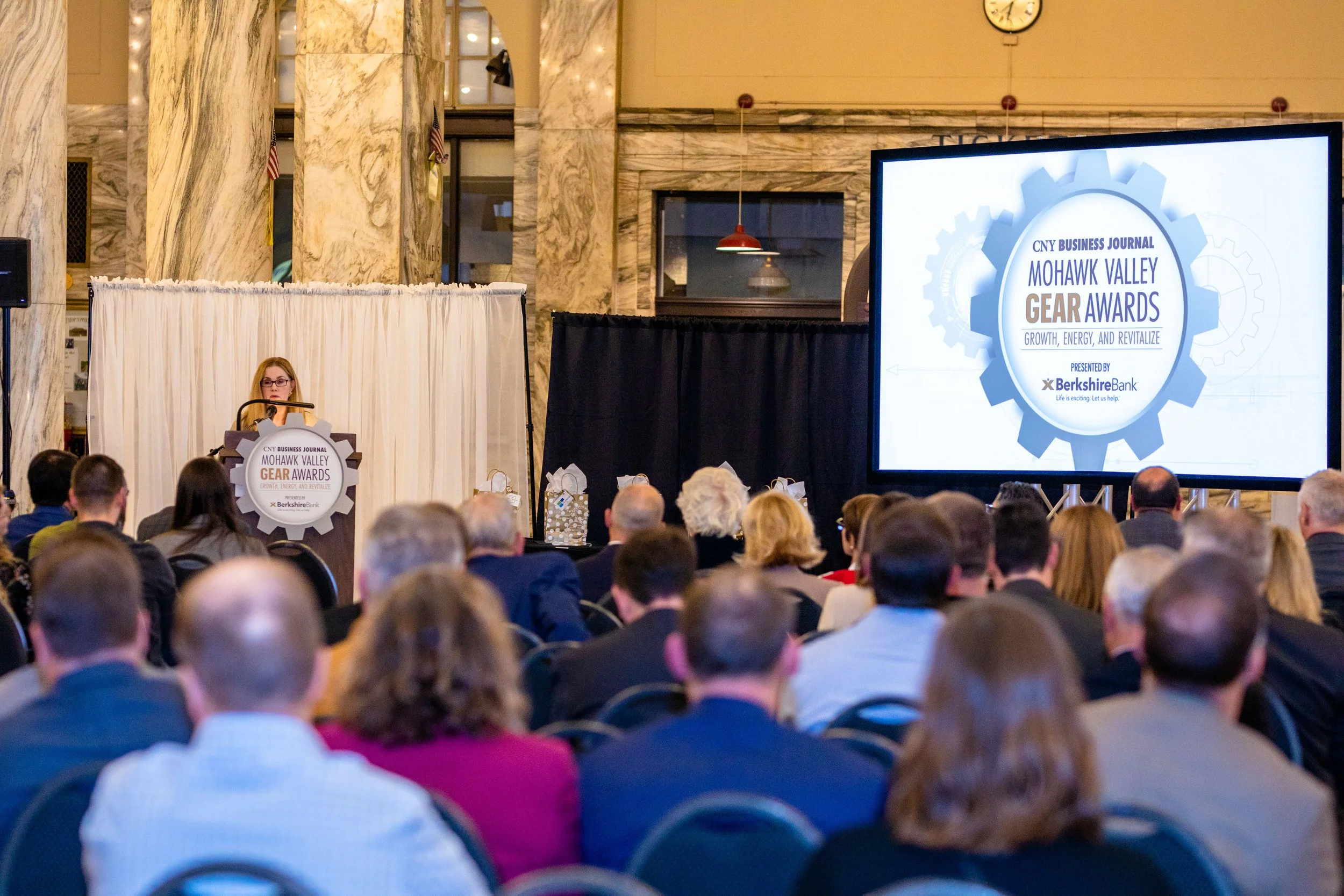 A woman speaking at a podium during an award ceremony with a large screen displaying the event's logo and title. The event is the Mohawk Valley Gear Awards, sponsored by Berkshire Bank, held in a grand hall with marble columns and a seated audience.