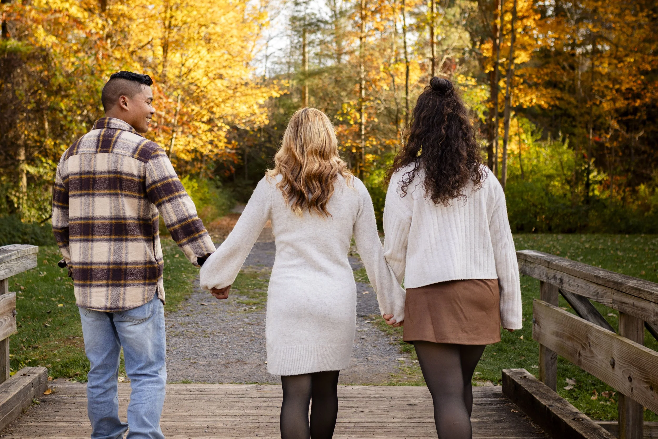 Three friends holding hands and walking on a wooden bridge through a park with fall foliage.