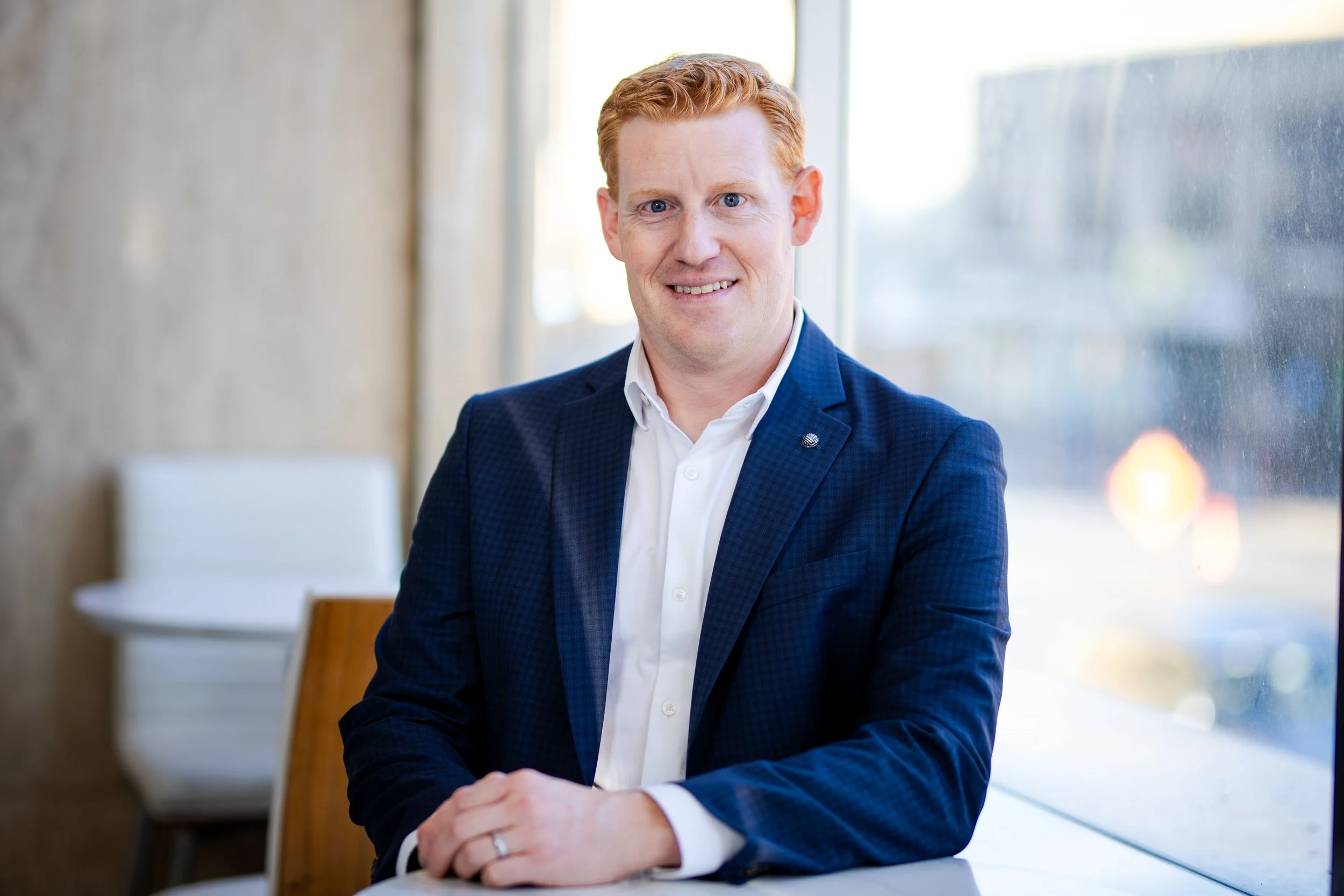 A man with red hair, wearing a white shirt and navy blue blazer, sitting at a wooden table near a window.