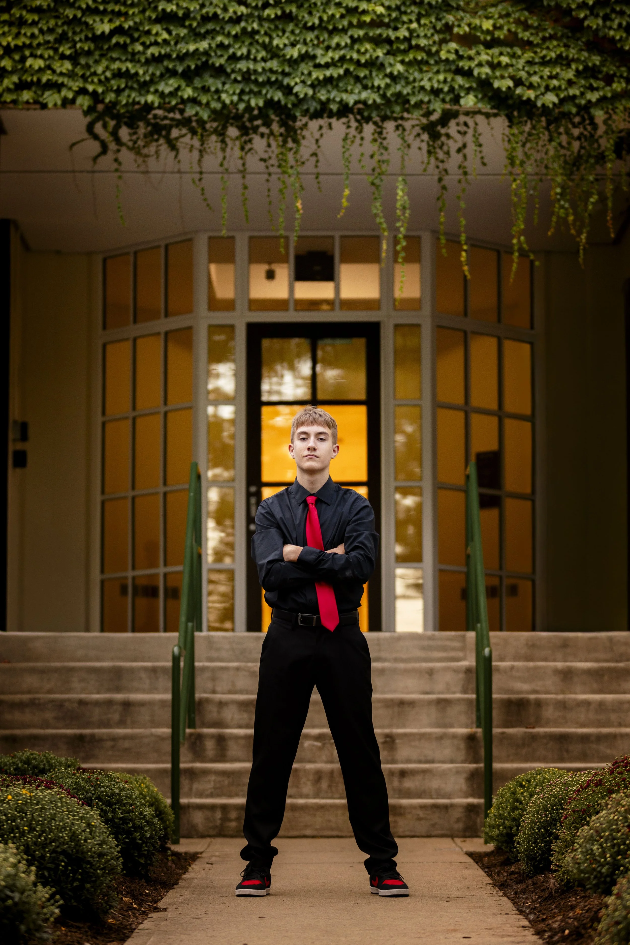 A young man in a black shirt, red tie, and black pants standing with arms crossed in front of a modern house with large glass windows and front steps, during sunset.