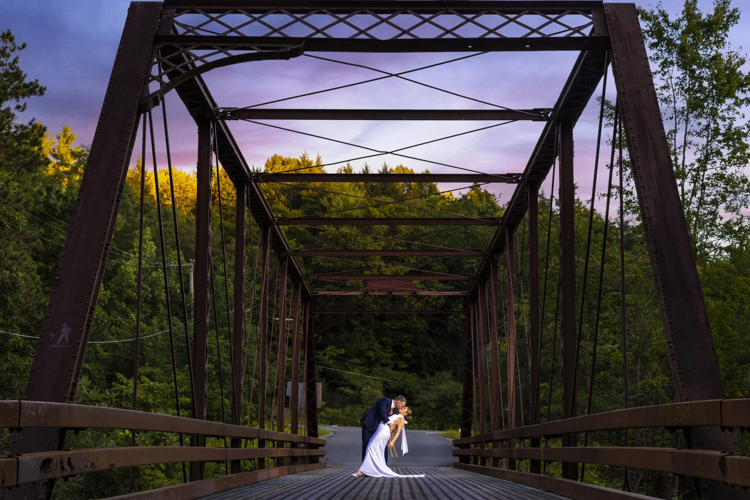 A couple getting married on a metal bridge surrounded by green trees at sunset.