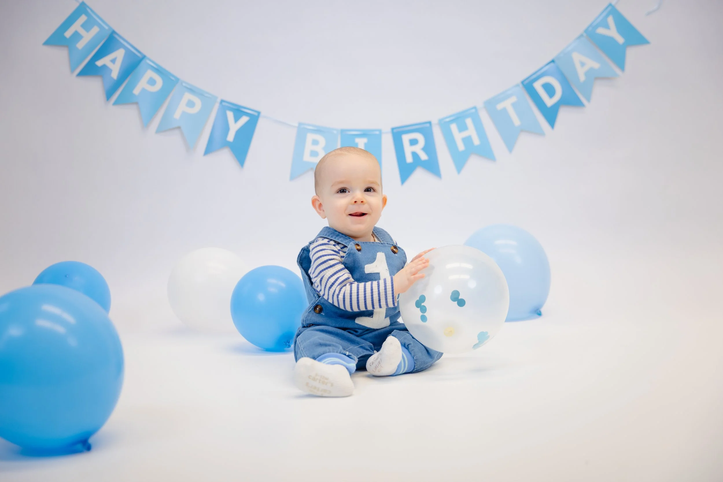 Baby sitting on white background with blue and white balloons, wearing a blue and white striped shirt and blue overalls, with a blue 'Happy Birthday' banner overhead.