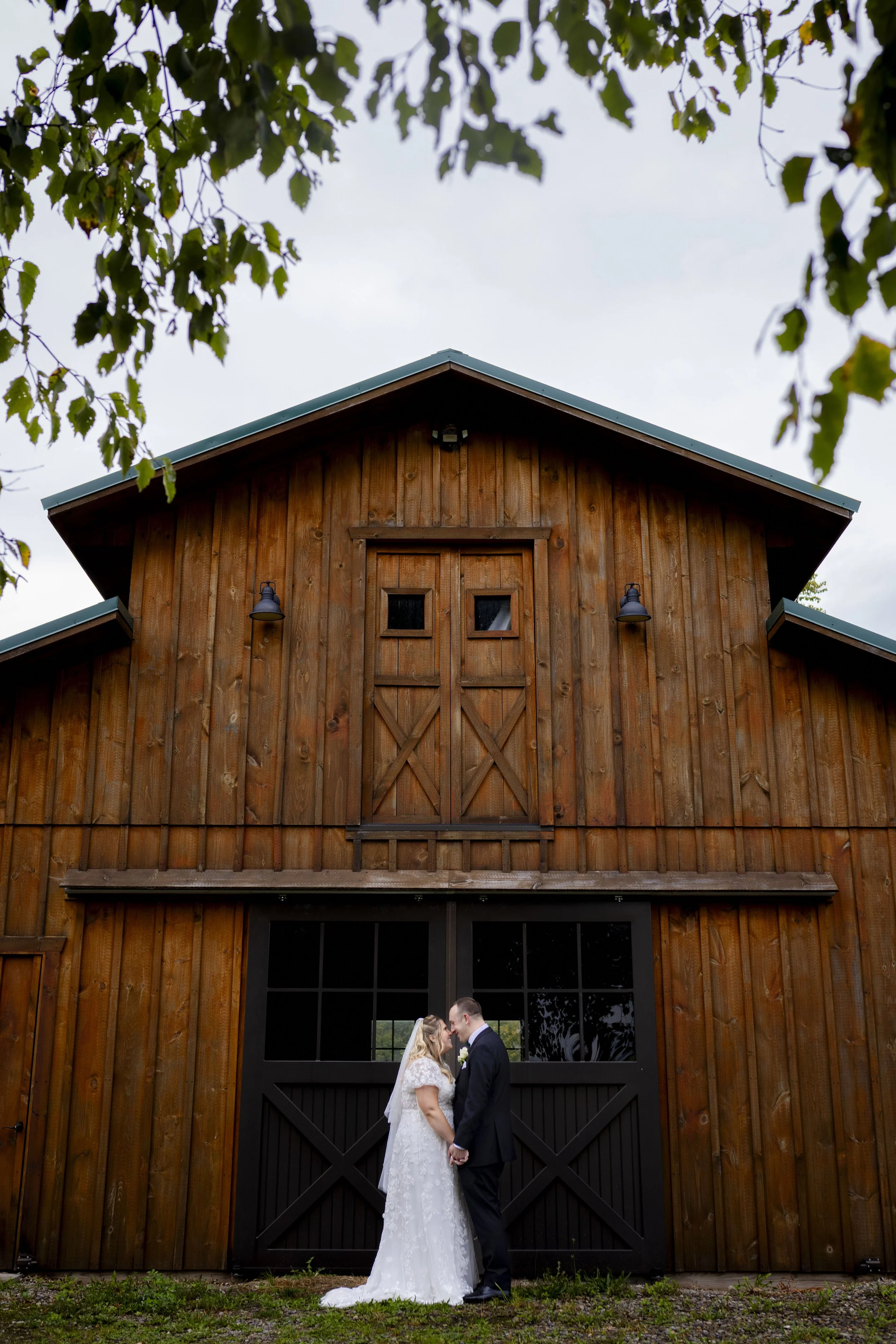A bride and groom standing close together in front of a large wooden barn, holding hands and touching foreheads, with trees and an overcast sky in the background.