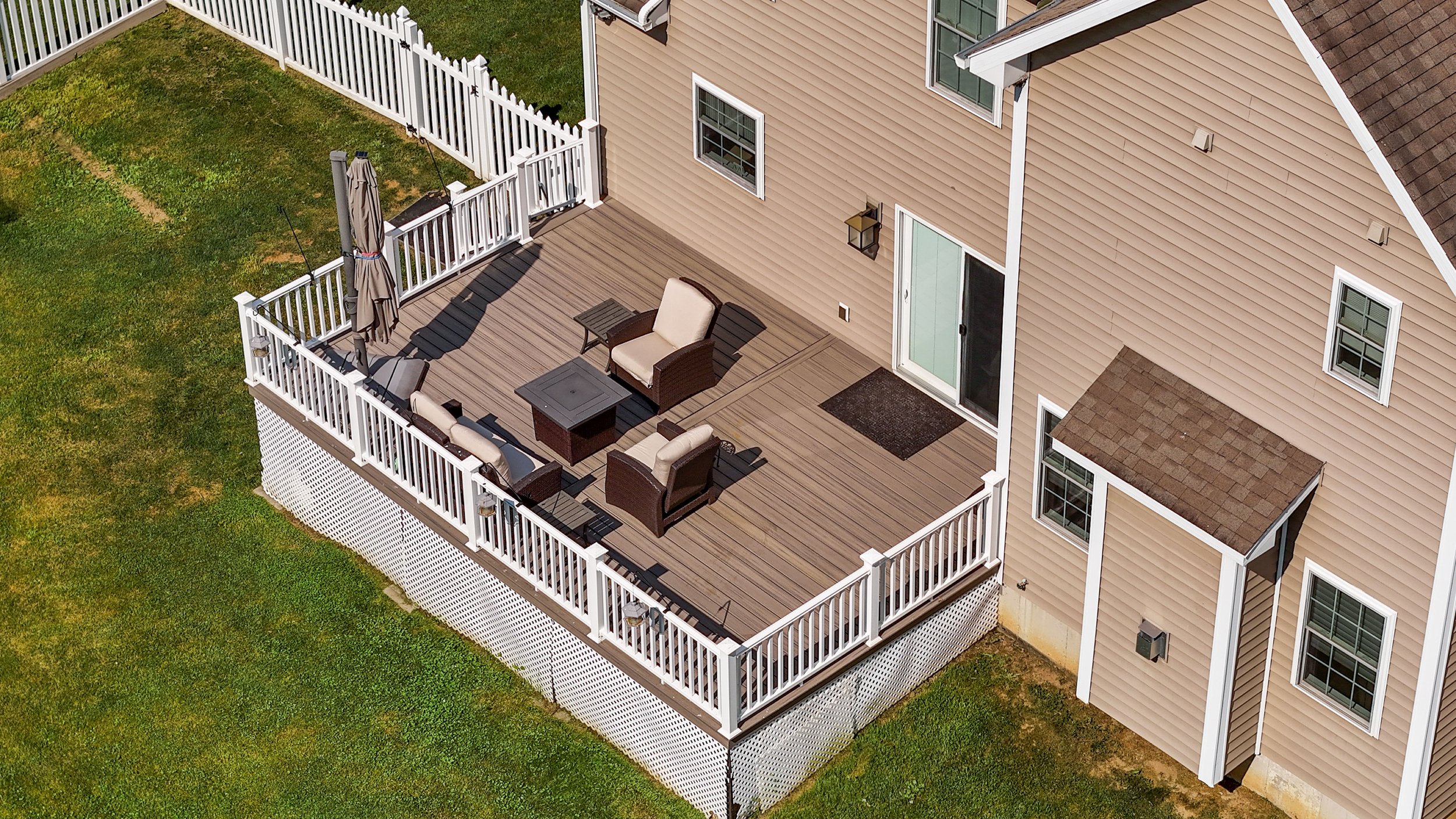 A large wooden deck attached to a beige house with white trim, featuring outdoor furniture including a sofa, chairs, tables, and a closed umbrella, surrounded by a white picket fence with grass yard beyond.