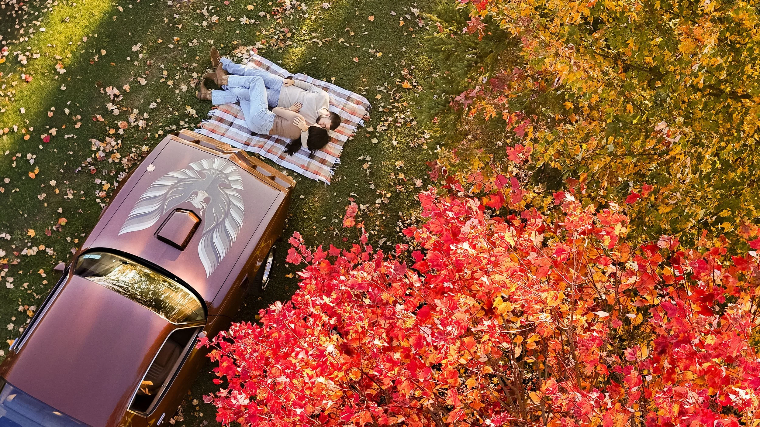 A couple lying on a plaid blanket on the grass next to a parked truck with a large lion head graphic on it, surrounded by fall foliage with red, orange, and yellow leaves.
