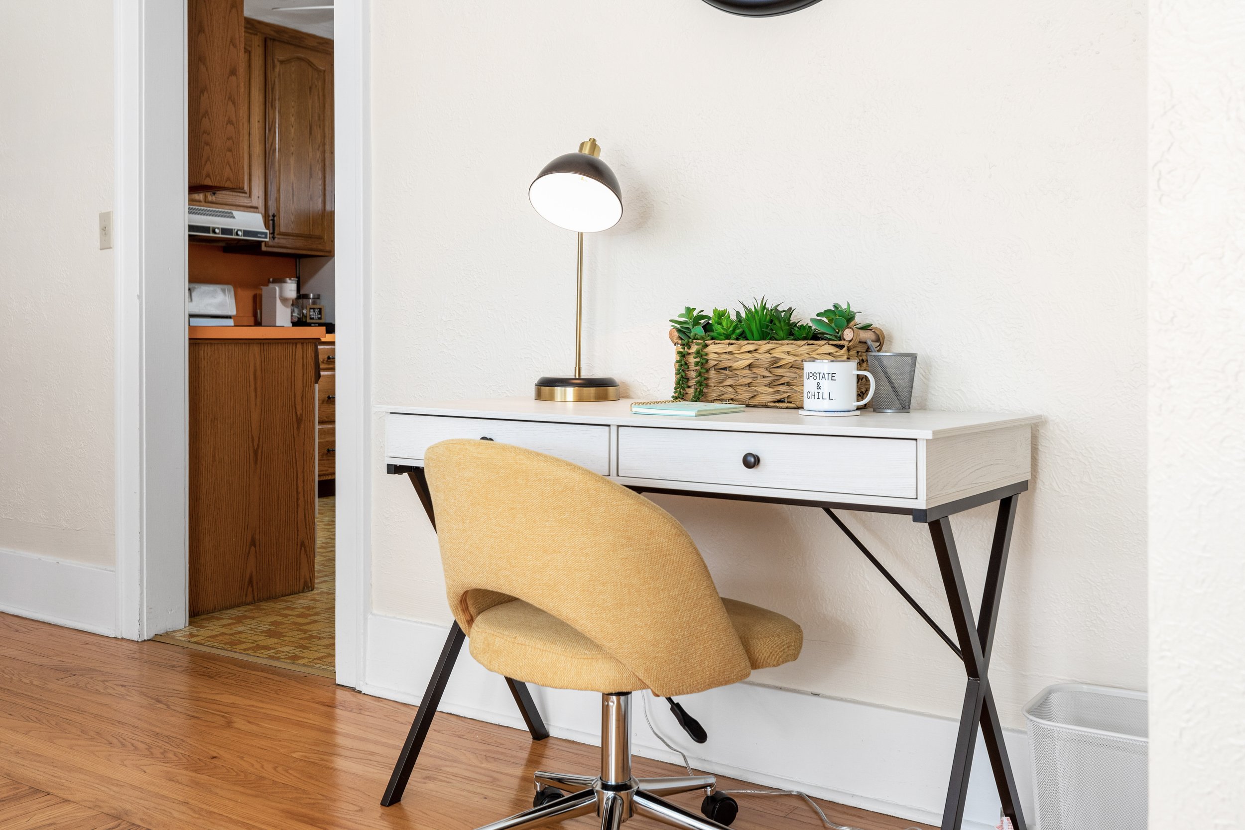 Home office area with a white desk, yellow office chair, lamp, and decor including a basket with green plants and a mug that says 'Upstate & Chill.'